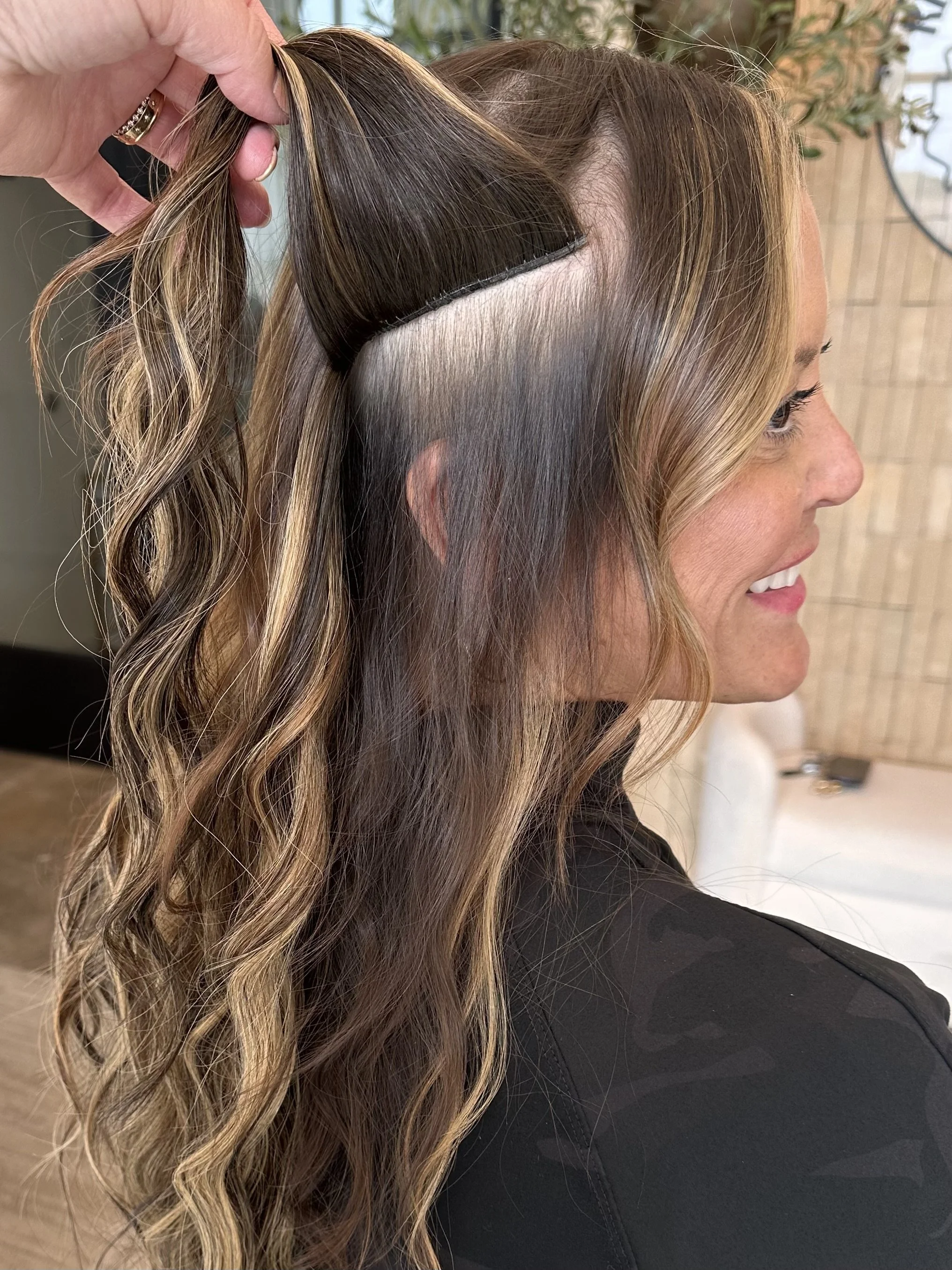 A woman with long, wavy, brown hair with blonde highlights is having her hair styled. A stylist is lifting a section of her hair to reveal a fresh undercut fade with short, shaved sides that transition into longer hair on top.