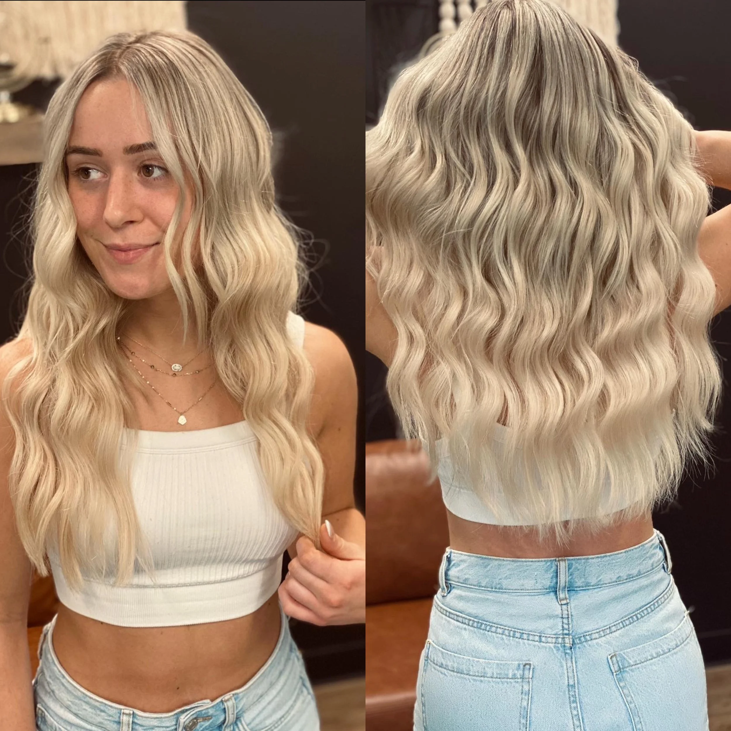 Woman with long, wavy blonde hair, wearing a white sleeveless crop top and light blue jeans, posing in front of a dark background.