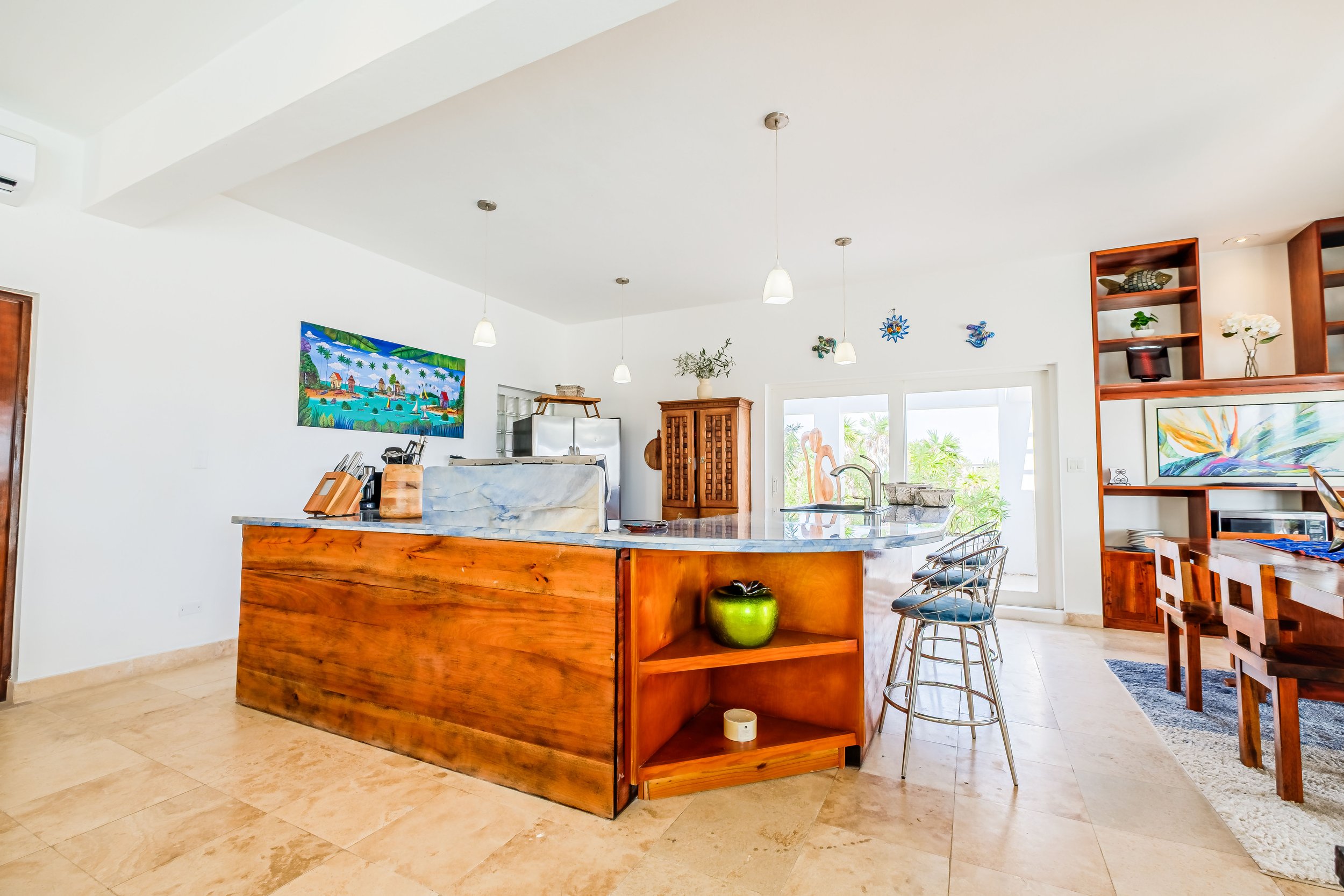 Modern kitchen with wooden cabinets, marble countertop, and bar stools. Features a colorful wall painting and decorative shelving. Bright and spacious with natural light from large windows.