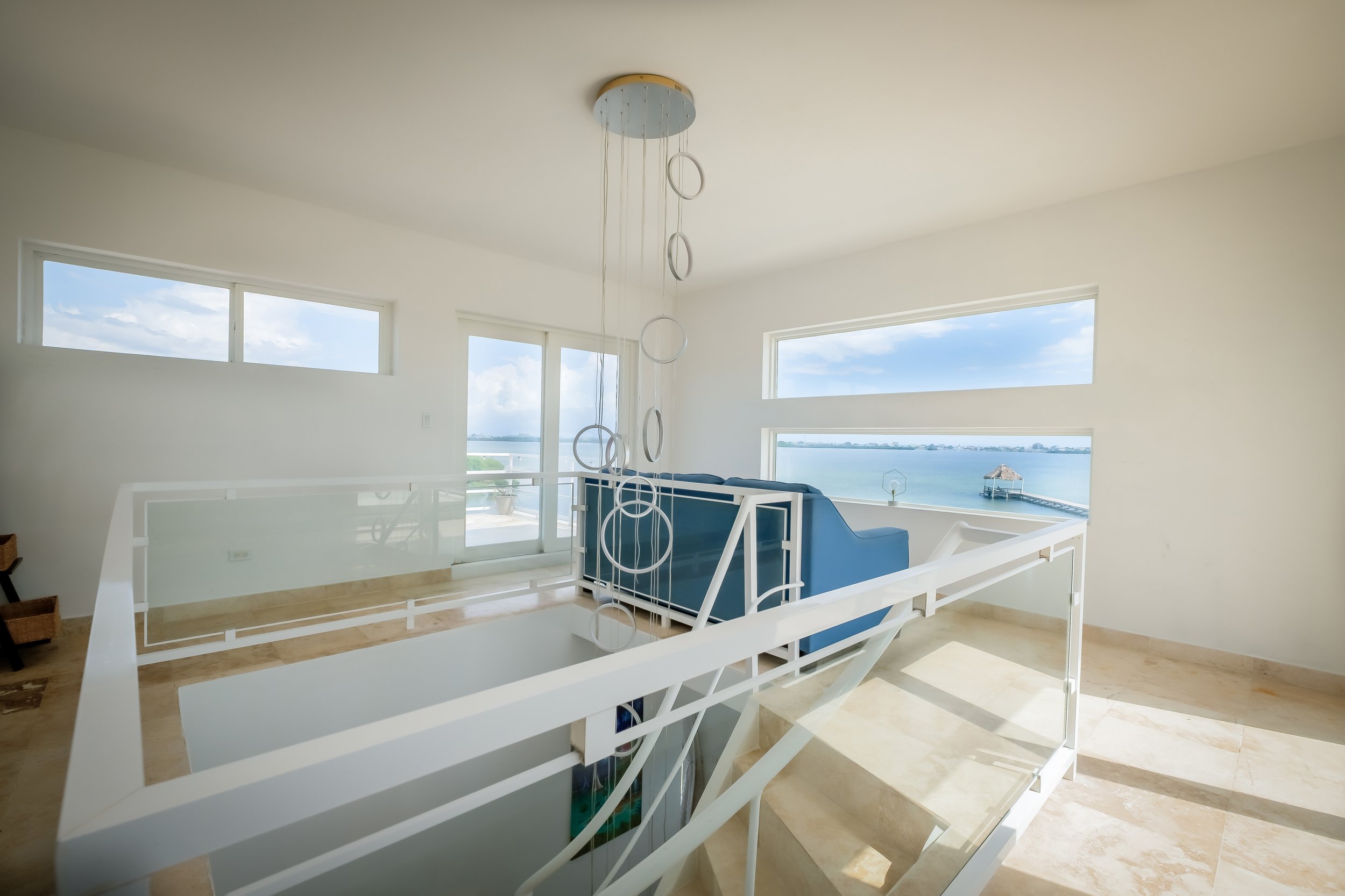 Modern interior with ocean view, featuring a staircase, large windows, a blue sofa, and a hanging light fixture.