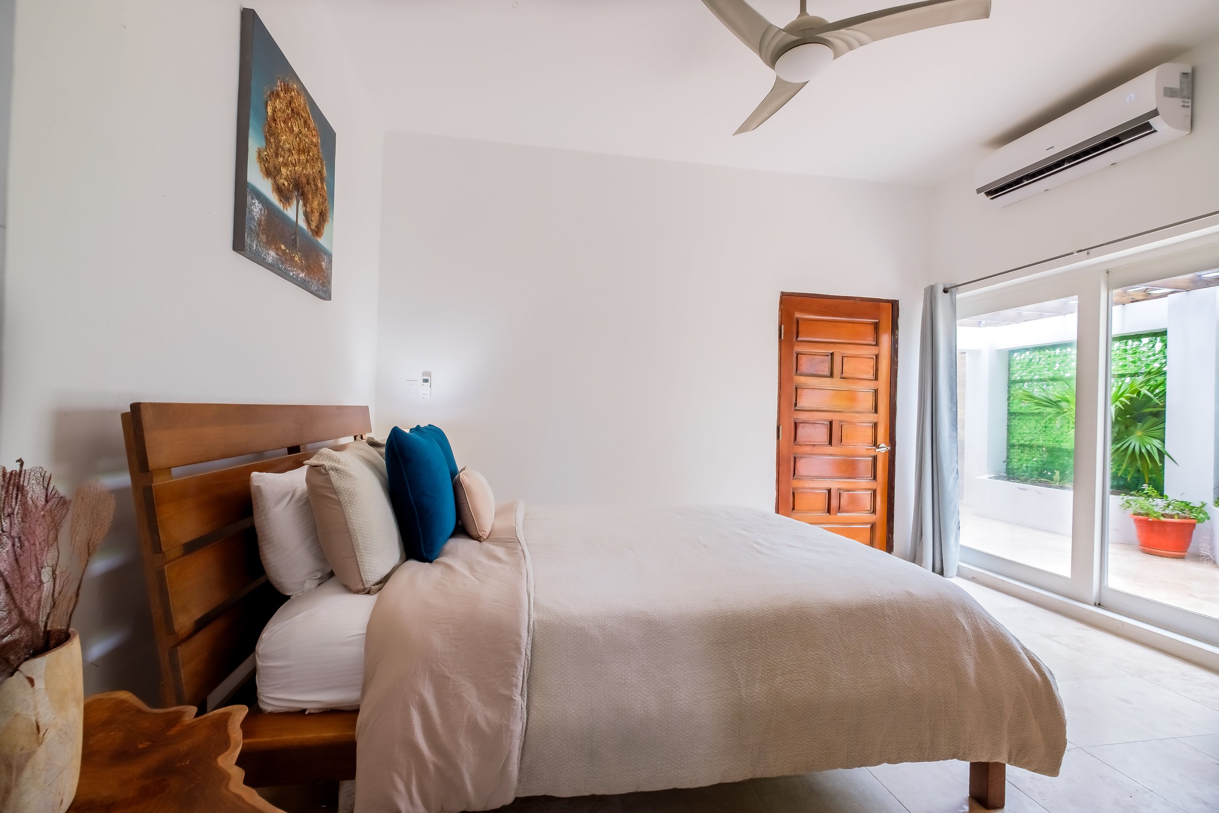 Modern bedroom with a wooden bed, white and blue pillows, beige bedding, wall art, ceiling fan, wooden door, air conditioner, and glass sliding doors leading to a patio with greenery.