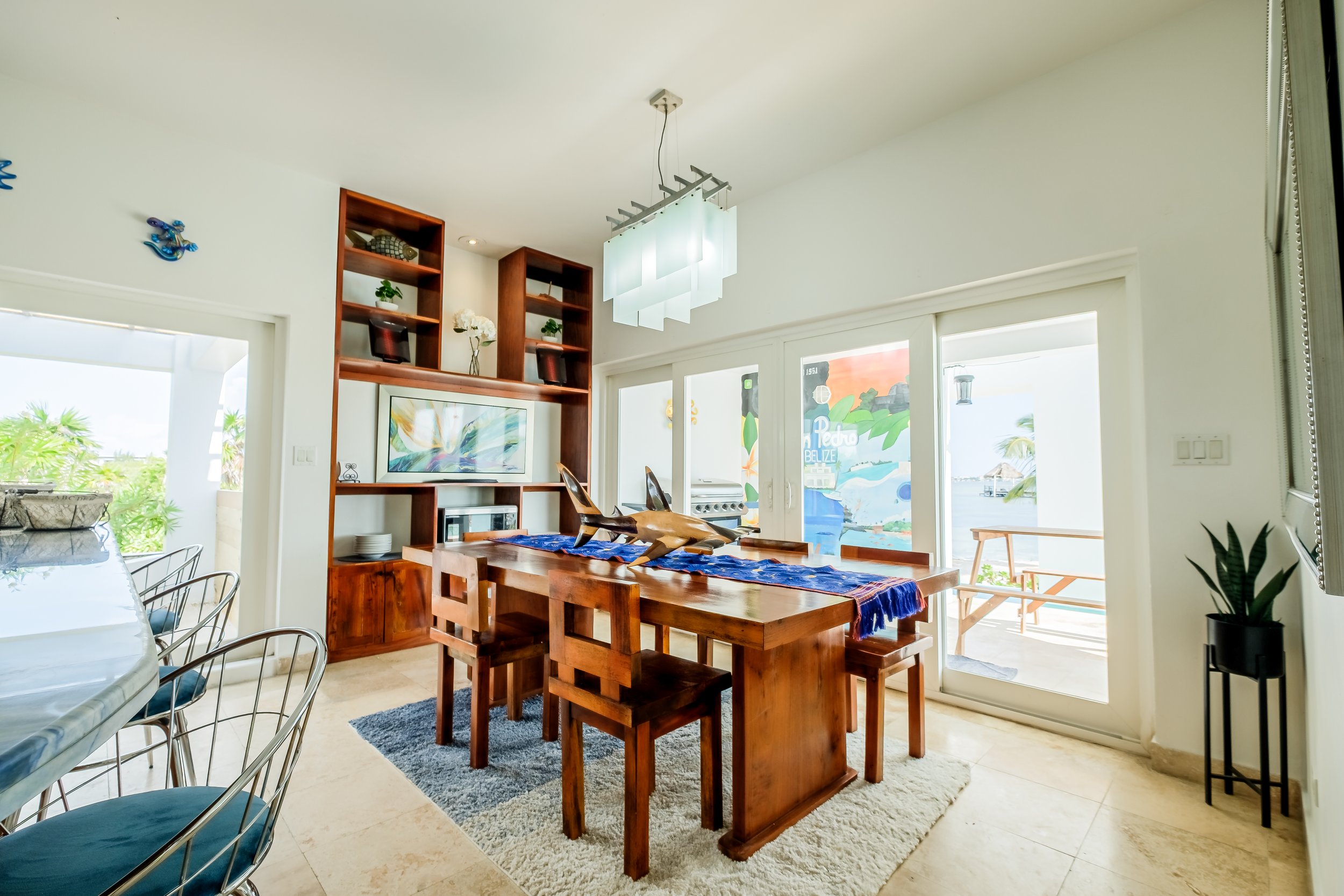 Modern dining room with wooden table and chairs, built-in shelving, decorative lighting, and large glass doors leading to a balcony with an ocean view.