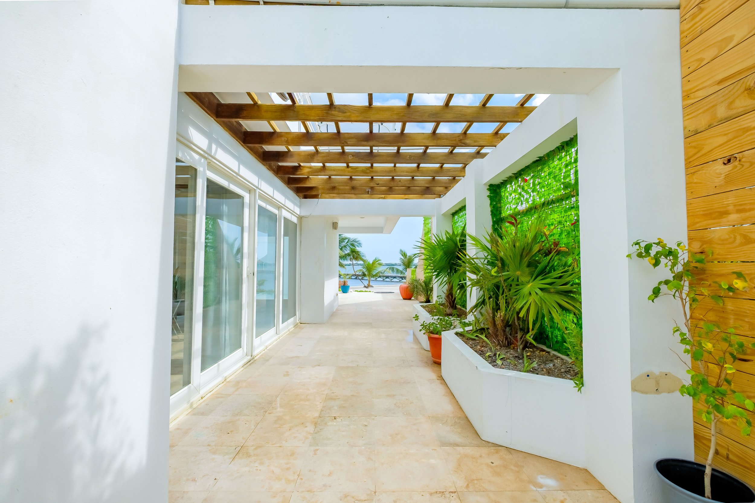 Outdoor hallway with wooden pergola, potted plants, white walls, glass doors, and a view of palm trees and the ocean in the background.