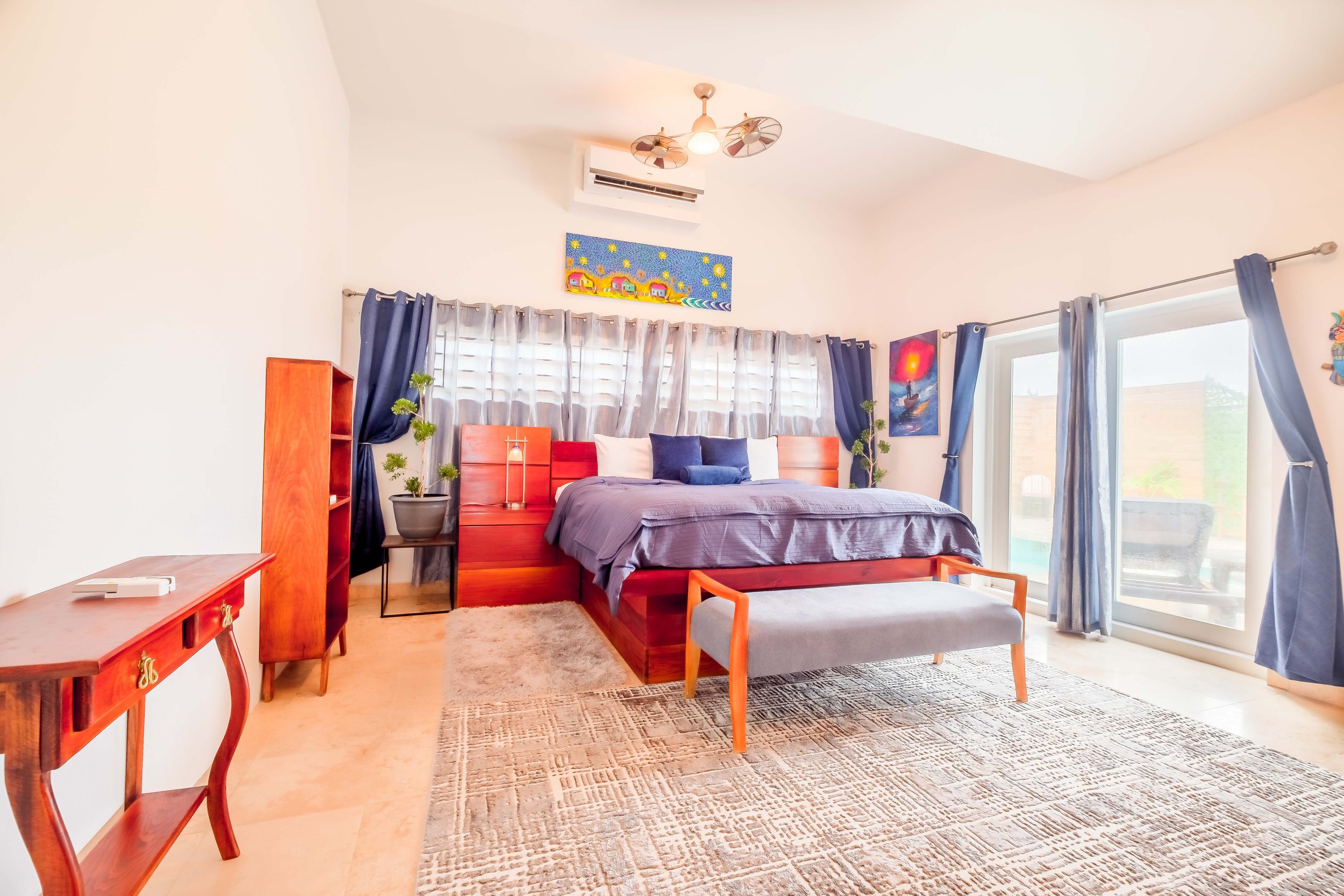 Modern bedroom with wooden furniture, a large bed with blue bedding, potted plants, wall art, and a sliding glass door leading outside.
