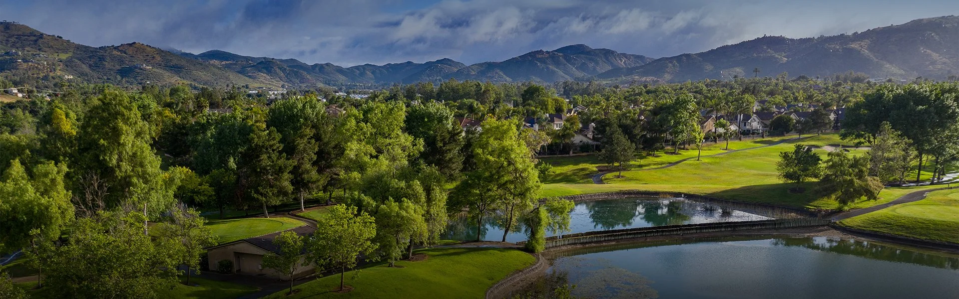 Scenic view of a golf course with ponds, surrounding trees, and houses, with mountains in the background under a partly cloudy sky.