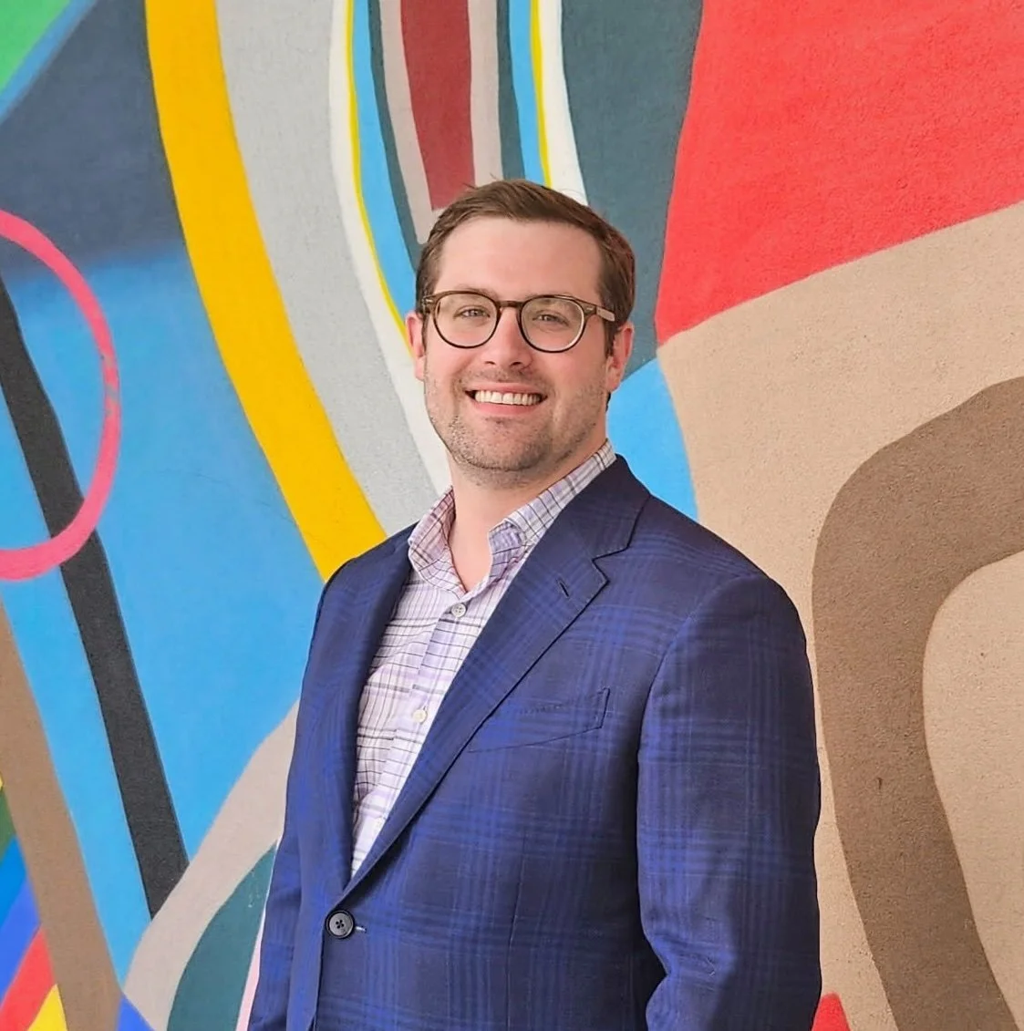 Smiling man in blue suit and glasses standing in front of colorful abstract mural.