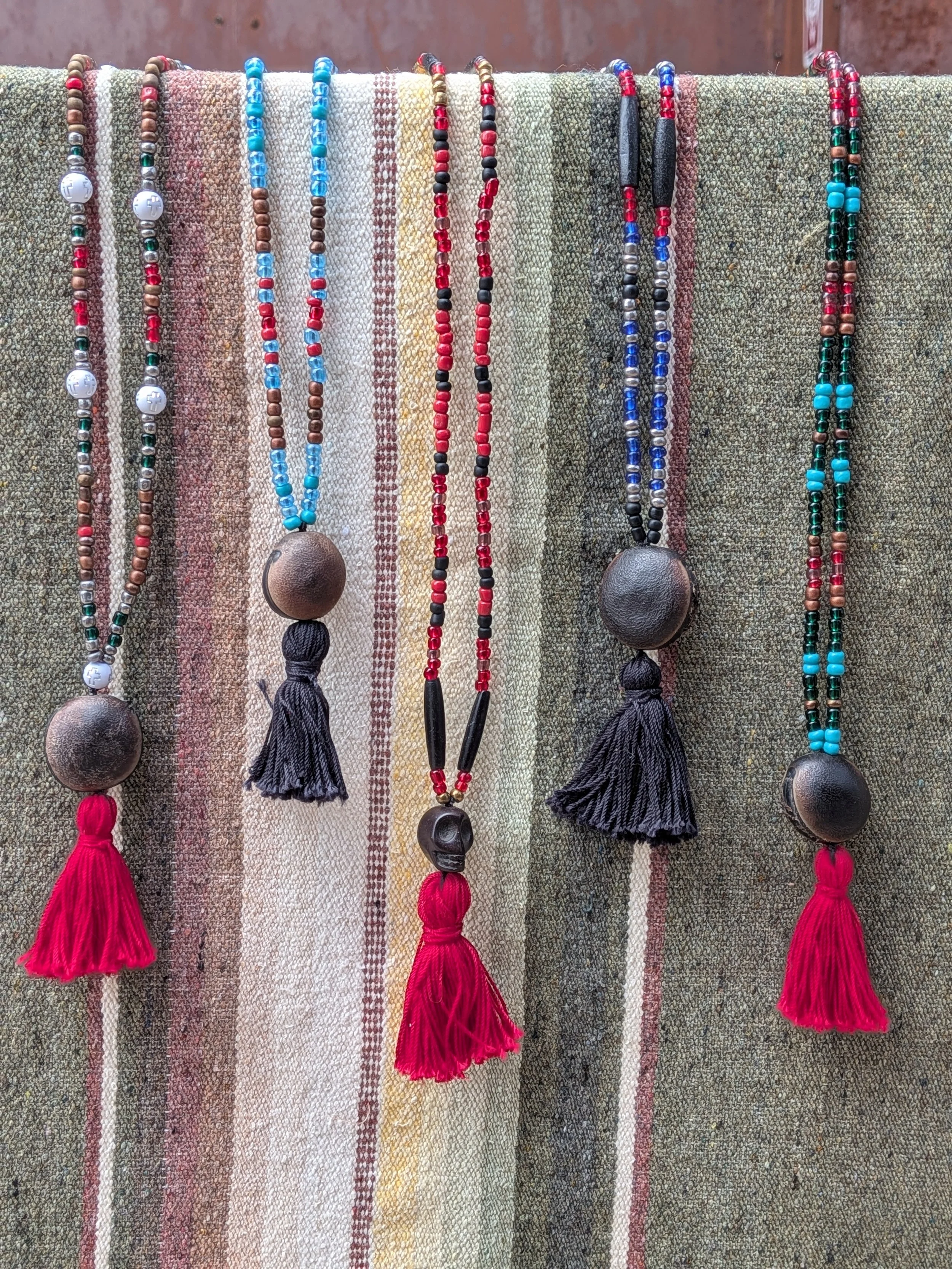 Fifteen beaded necklaces with tassels are displayed hanging against a colorful woven fabric background. The necklaces feature various bead colors including red, black, blue, turquoise, and metallic, with some having large round wooden or ceramic pendants and black tassels at the end.