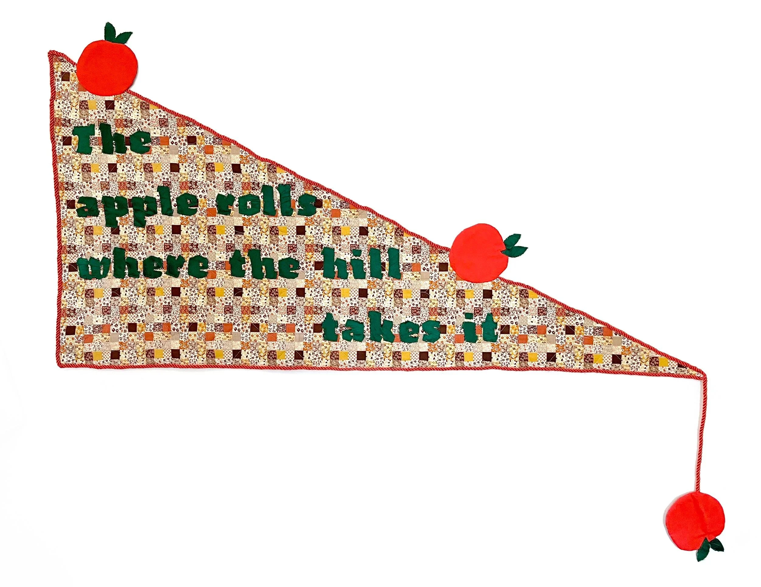 Triangle fabric quilt with applique that says "The apple rolls where the hill takes it". 3 apples look like they are in motion rolling down and off the triangle. It's shaped like a medieval banner with a plaid background, red trim, and green text.