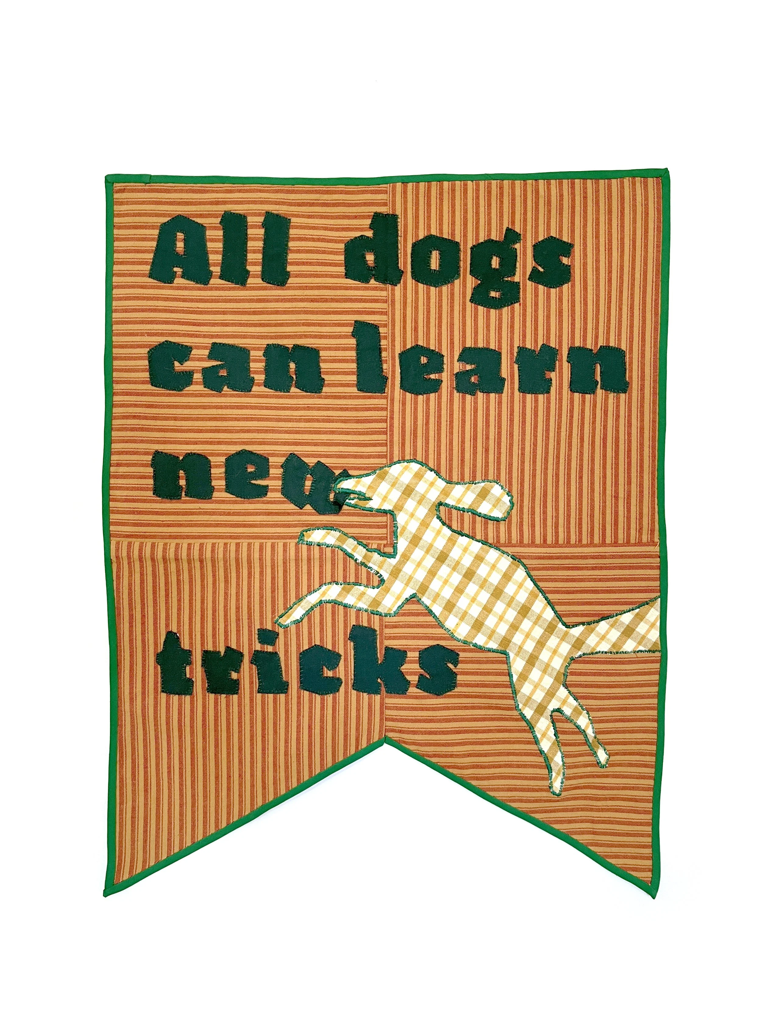 Fabric quilt with applique that says "All dogs can learn new tricks". A jumping dog is biting the letter "W". It is shaped like a medieval banner with red and yellow striped ground, green trim, and green text.
