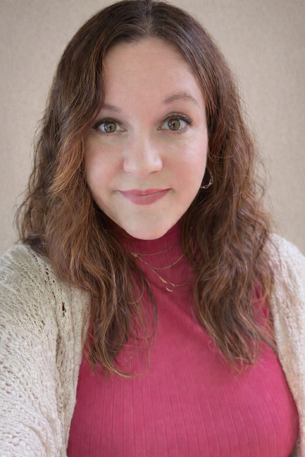 Close-up of a woman with wavy, shoulder-length brown hair and green eyes, wearing a red turtleneck, a beige knit cardigan, and a gold chain necklace, smiling softly at the camera with a neutral background.