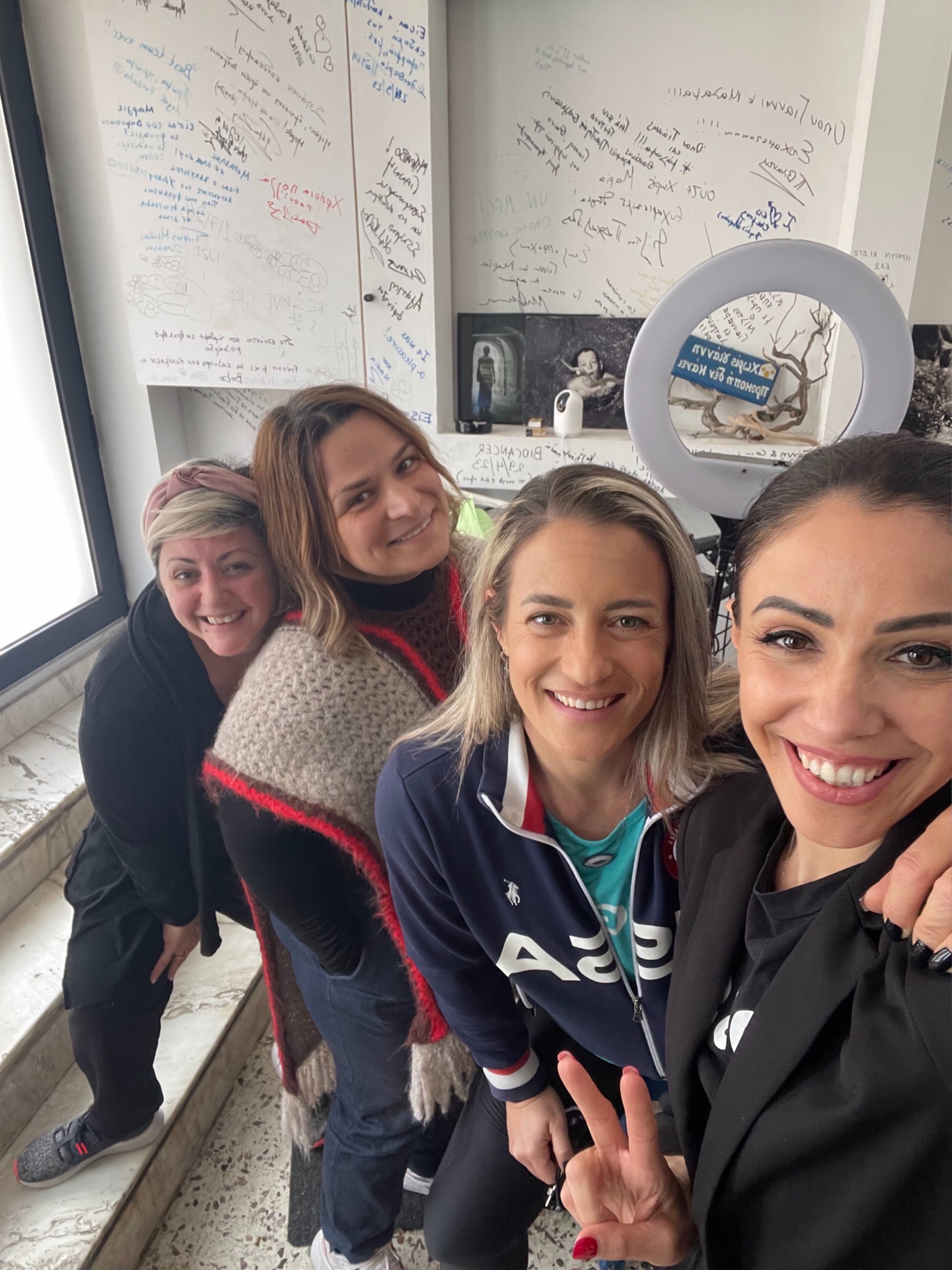 Four women smiling for a selfie in front of a wall covered with handwritten notes and drawings. One woman is making a peace sign. There is a circular ring light above them.