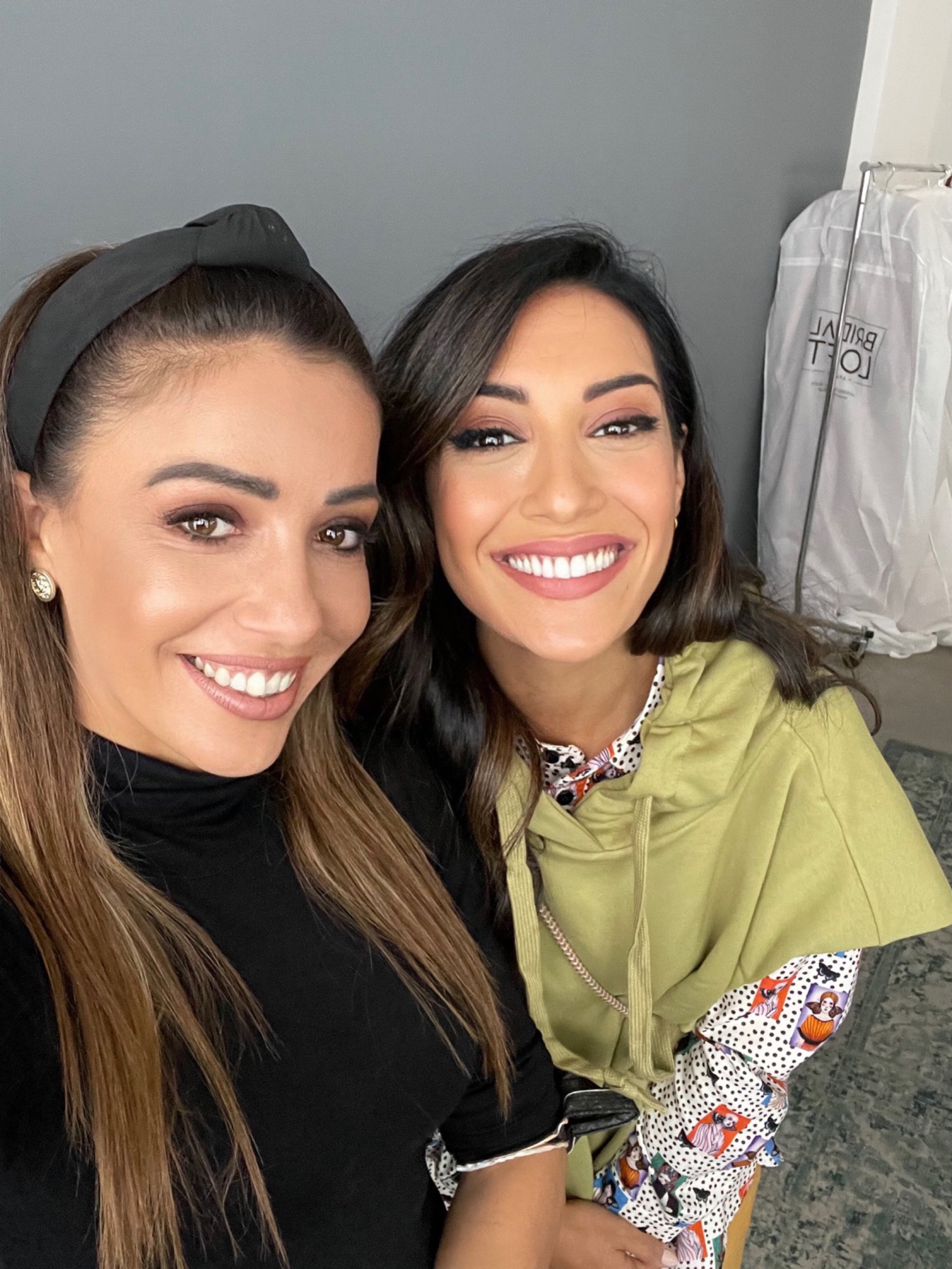 Two women smiling and taking a selfie indoors, with gray wall and white plastic-covered object in the background.