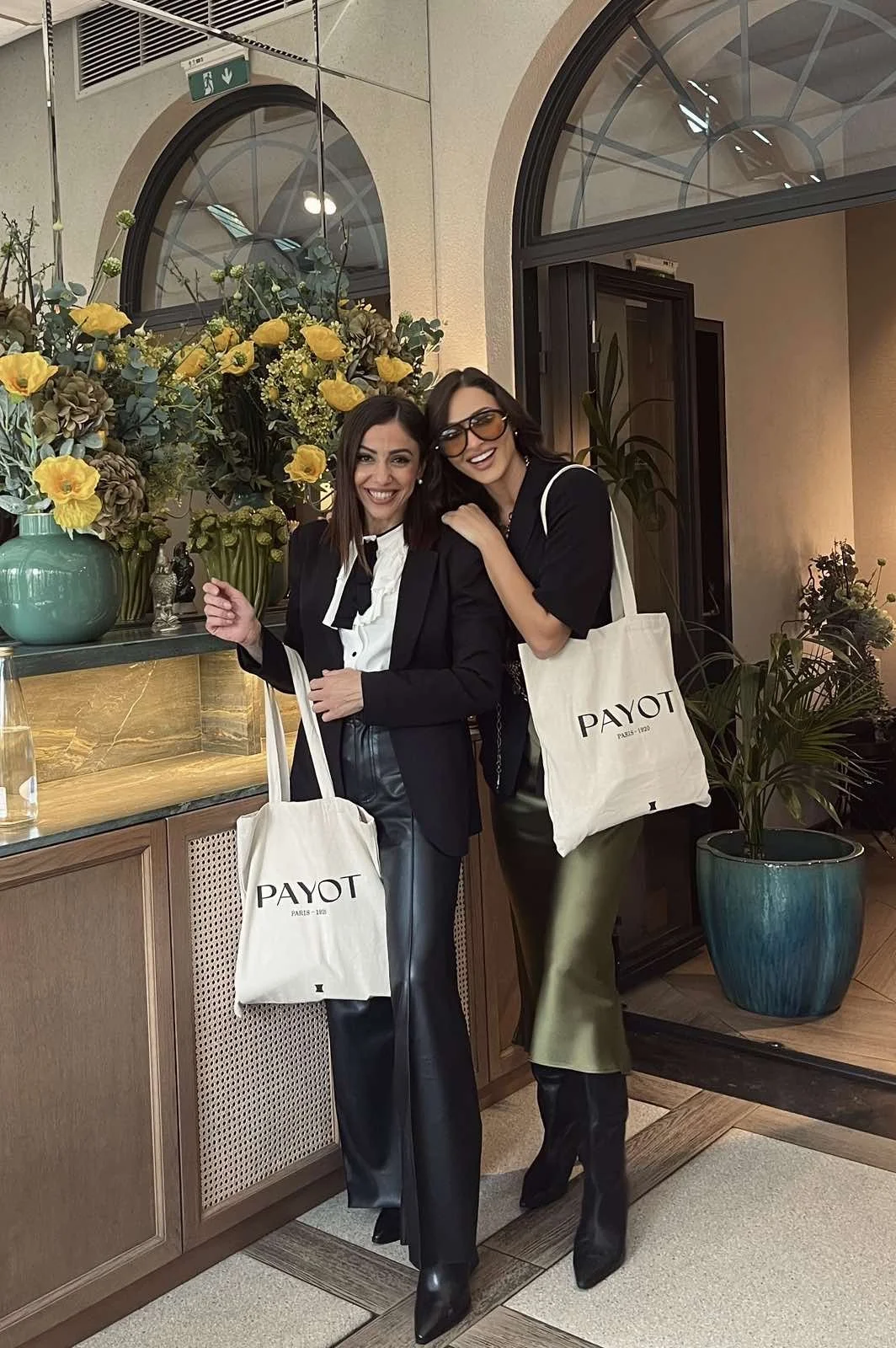 Two women smiling indoors, standing next to a floral arrangement and a large window. They are wearing black jackets, with one in leather pants and the other in a satin skirt. Both carry tote bags with the word 'PAYOT' printed on them.