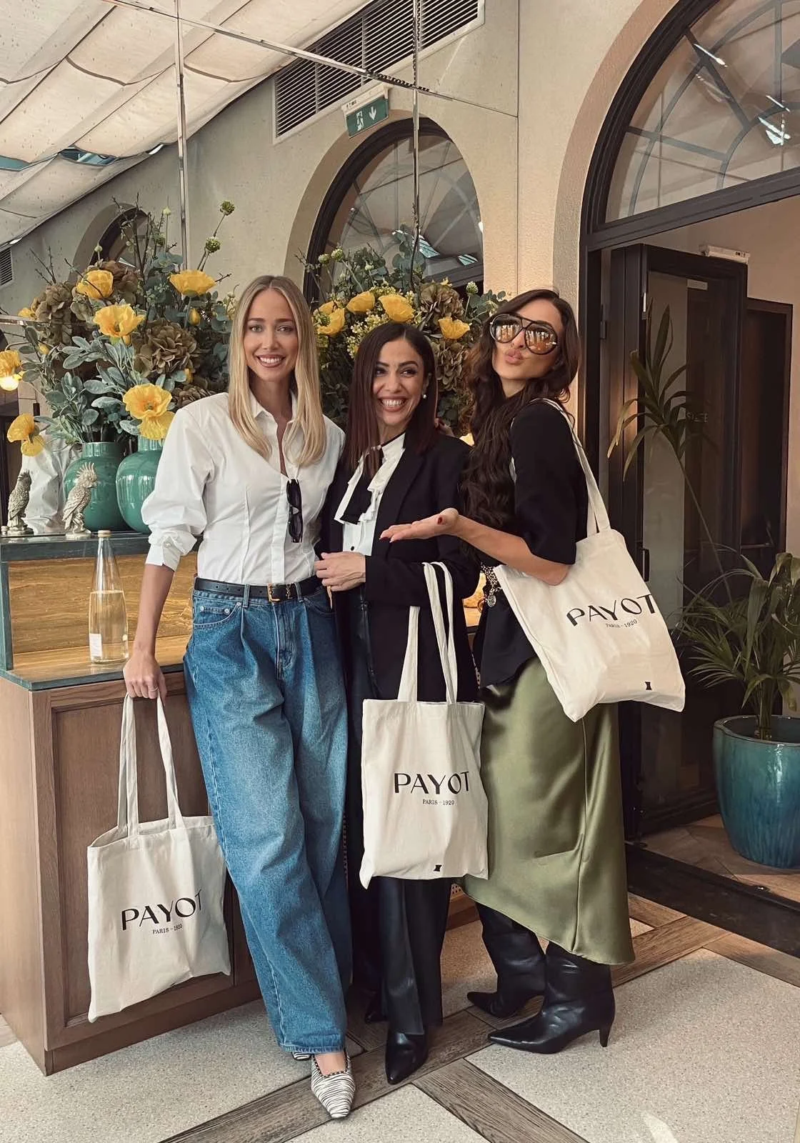 Three women smiling and posing together indoors in front of floral arrangements, holding white tote bags with the logo 'PAYOT'.