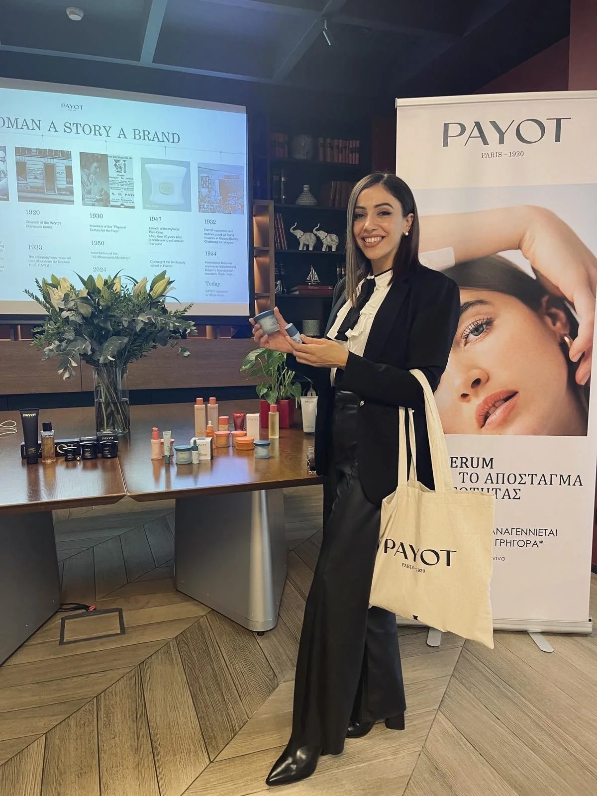 A woman standing at a presentation table with skincare products, holding a tube, smiling near a large banner with the brand name PAYOT and an image of a woman with skincare. A large screen behind her shows a timeline of PAYOT's history.