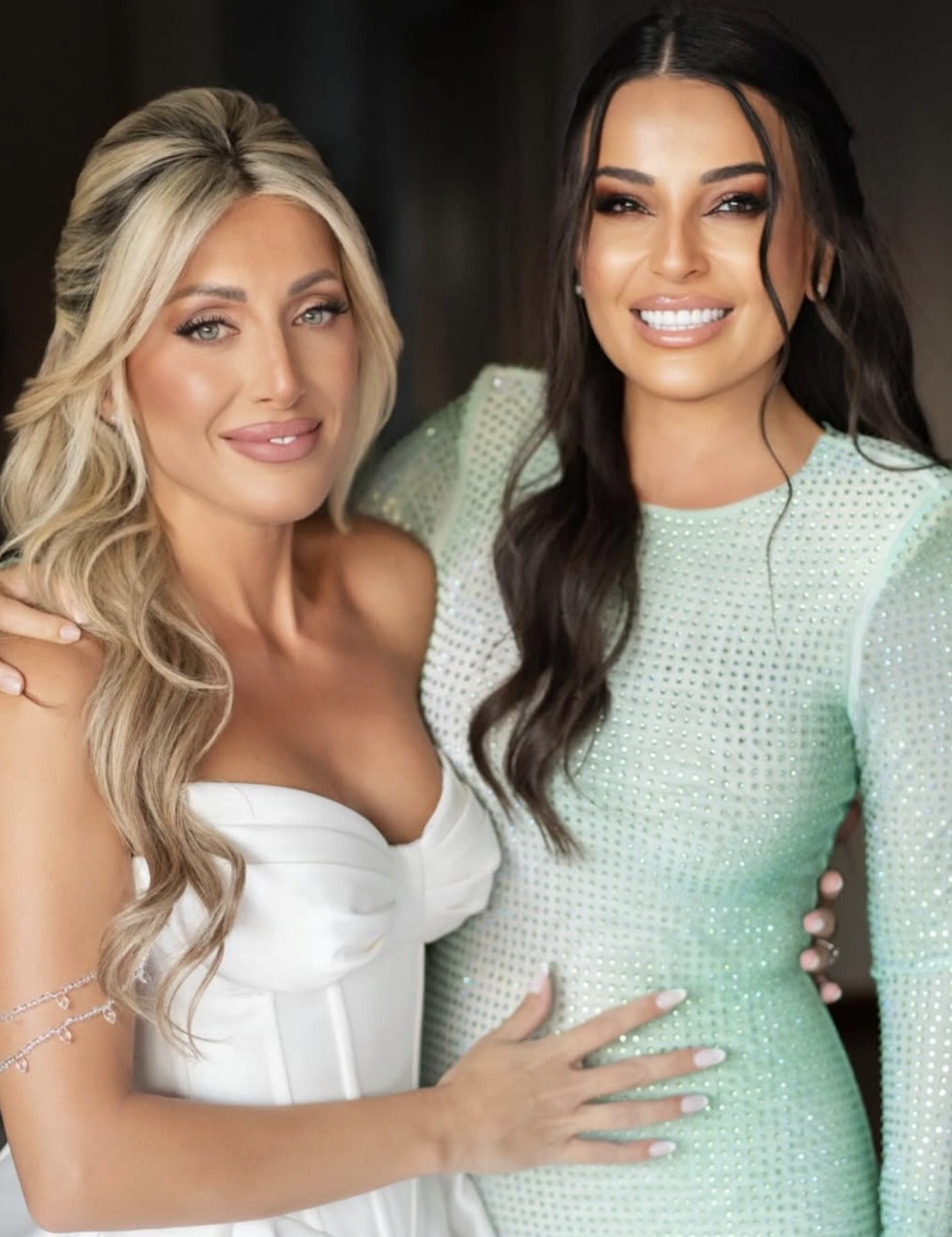 Two women smiling, one with blonde hair wearing a white dress, and the other with long dark hair wearing a light green dress with embellishments.