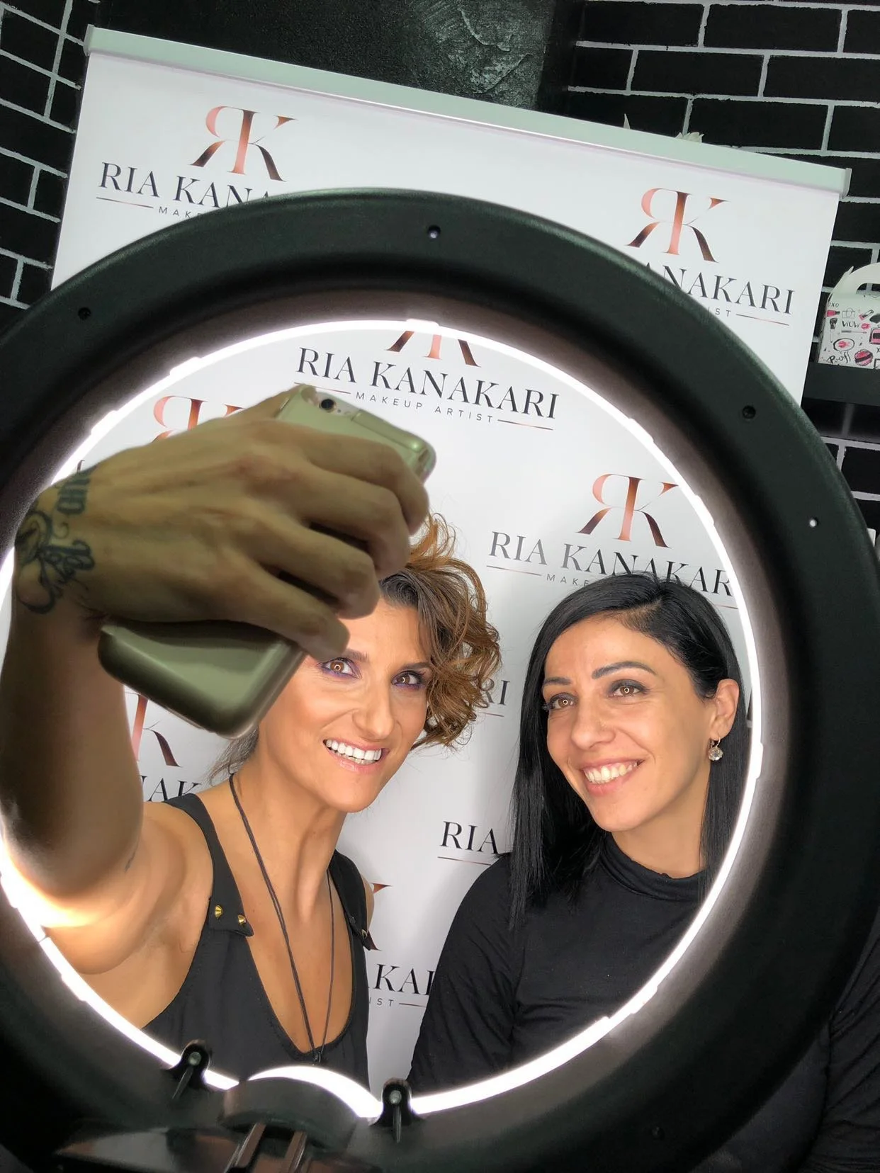 Two women taking a selfie in a ring light at a makeup studio, with a branded backdrop behind them that says 'Ria Kanakari Makeup Artist.'