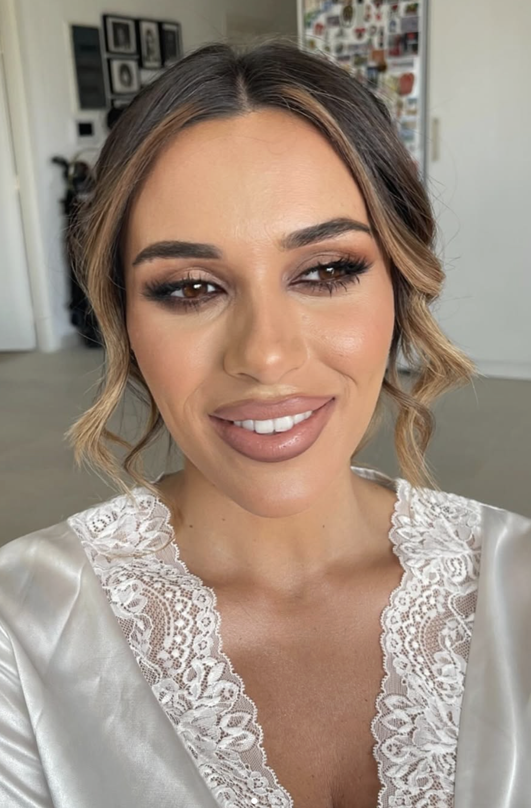 Close-up portrait of a woman with light skin, wavy brown hair with blonde highlights, wearing makeup with nude lipstick and eye makeup. She is smiling and wearing a satin top with lace trim. The background shows a wall with pictures and a locker.