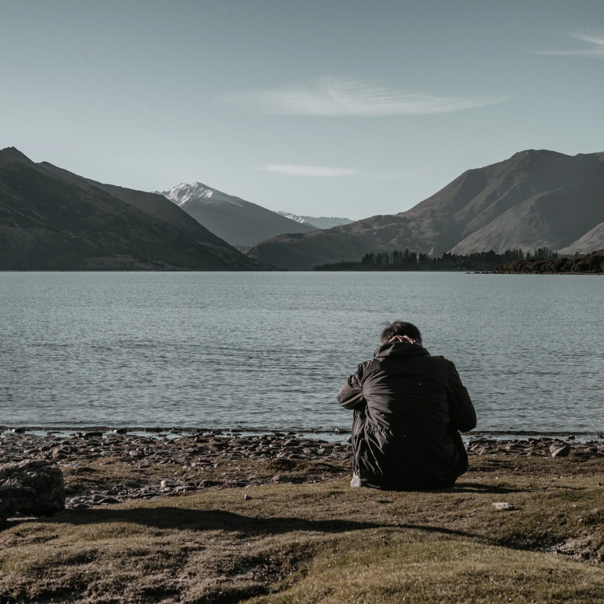A person sitting on the shore of a lake or river, looking at mountains in the distance under a cloudy sky.