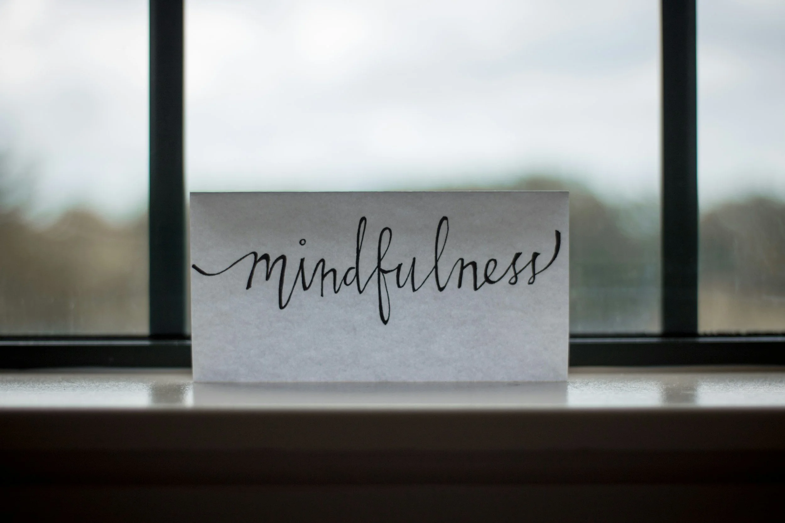 A handwritten sign on paper with the word 'mindfulness' placed on a windowsill, with blurred outdoor scenery visible through the window.
