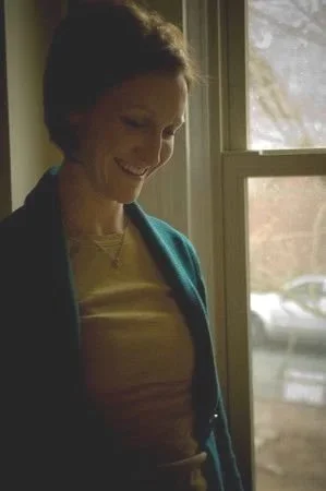 A woman smiling while looking down, standing near a window with sunlight coming through.