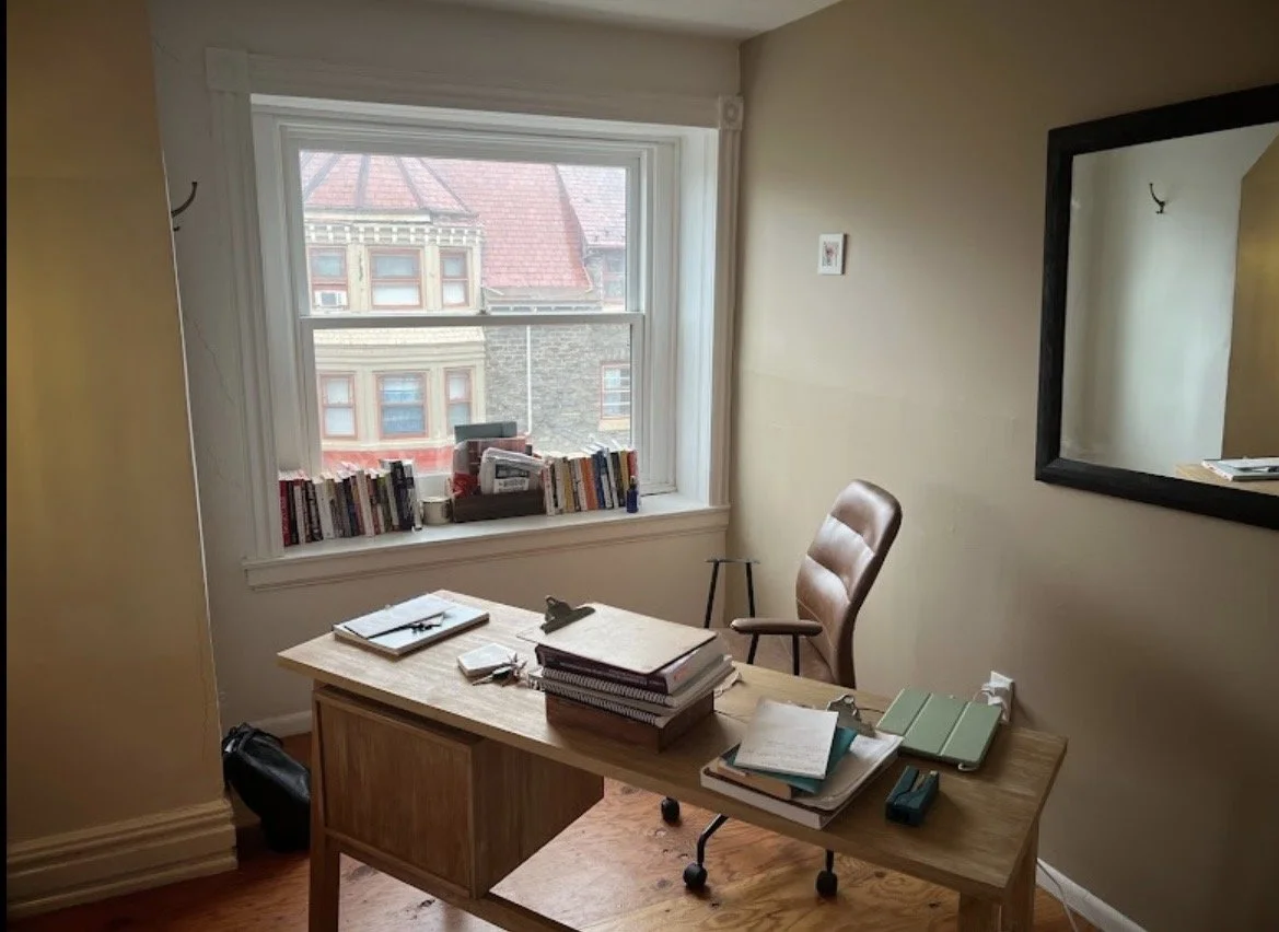 Home office with a wooden desk, stacked notebooks and papers, brown leather chair, window with books on the sill, and a large mirror on the wall.