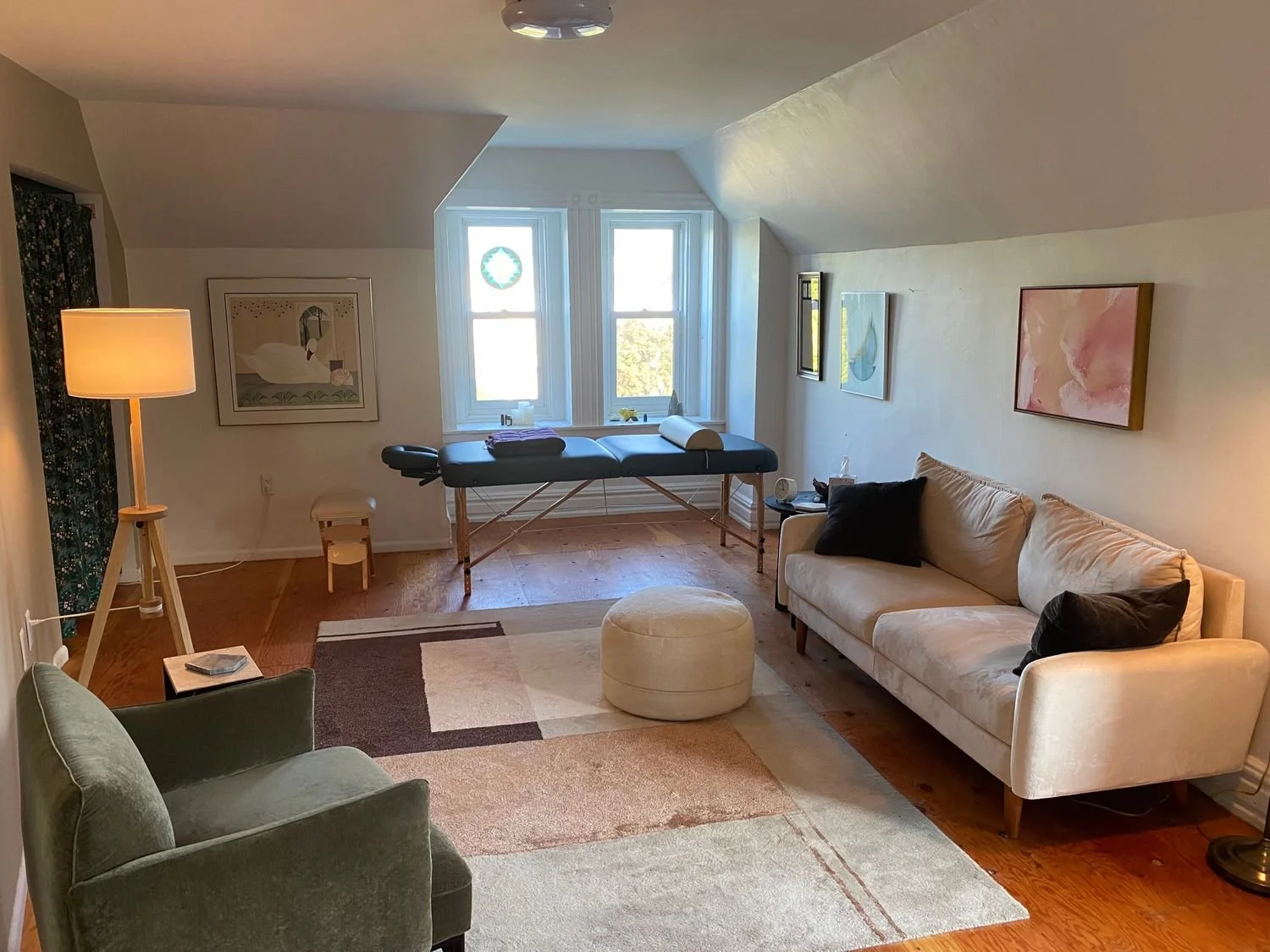Living room with a beige sofa, green armchair, patterned area rug, standing lamp, massage table near windows, abstract paintings on the walls, window with a wreath, and small decorative items.