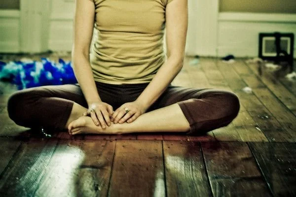 A person sitting cross-legged on a wooden floor, wearing a beige top and dark pants.
