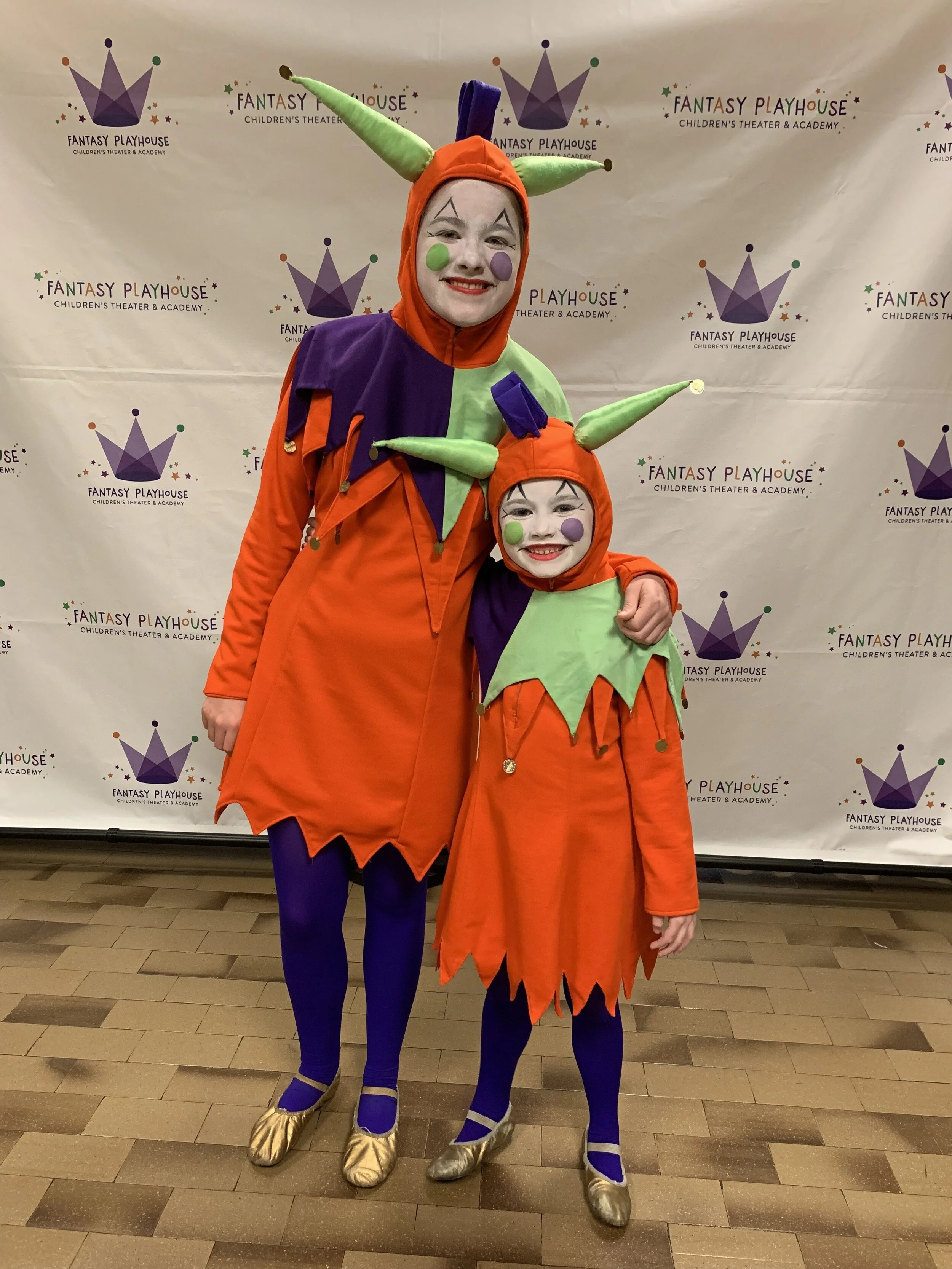Image of an adult and child in orange, purple and green jester costumes. This is Fantasy and Fantasy Jr.