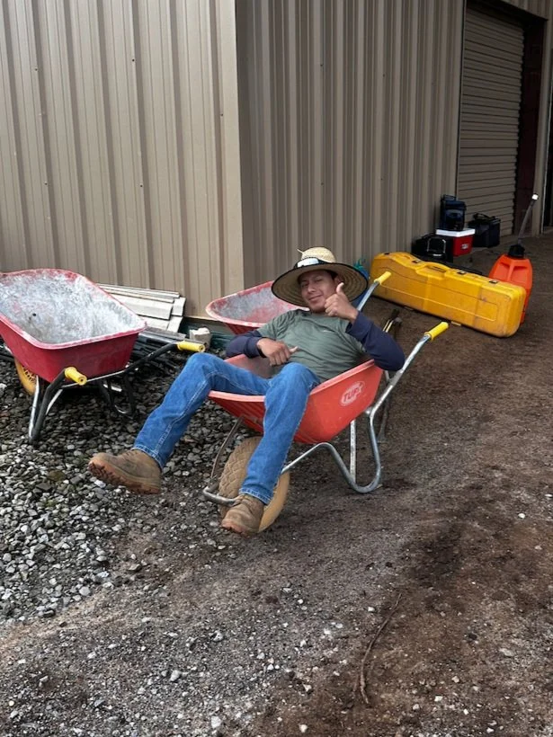 A person sitting in a red wheelbarrow, smiling and giving a thumbs-up, wearing a large sun hat, jeans, and work boots, with various tools and equipment in the background.