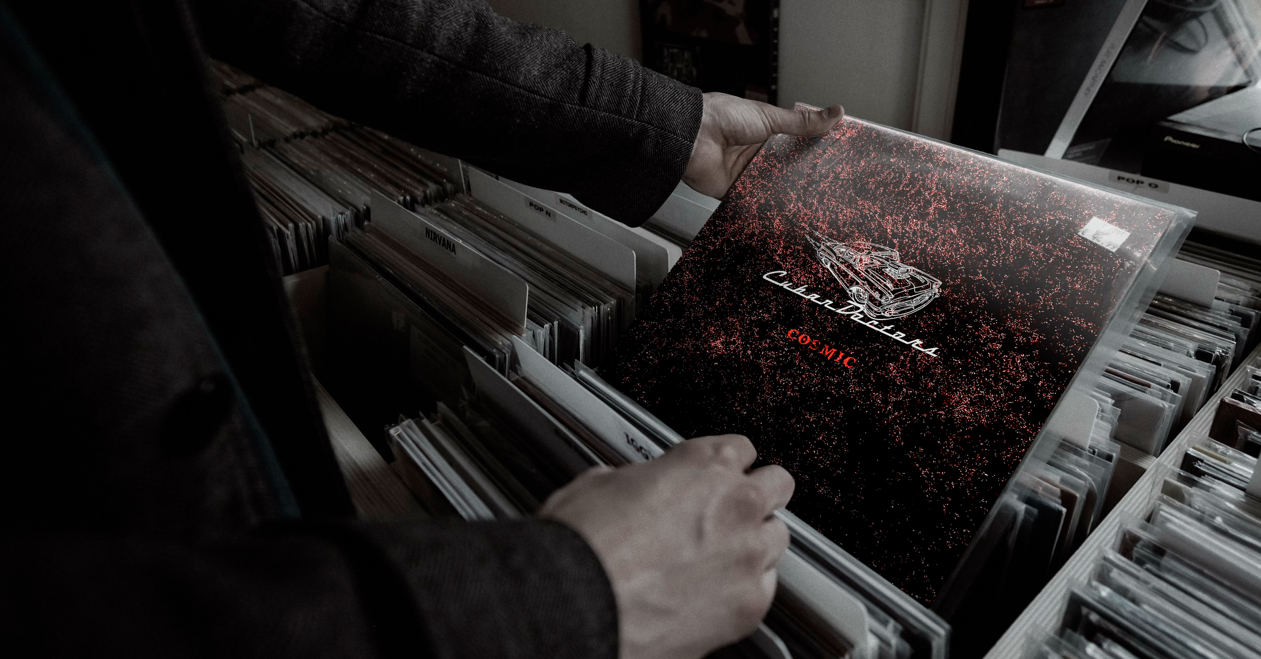Person browsing vinyl records in a store, holding an album with a black cover featuring a car illustration and the text 'Cosmic' and 'Cadillac'. The store has shelves filled with records labeled with artist names.