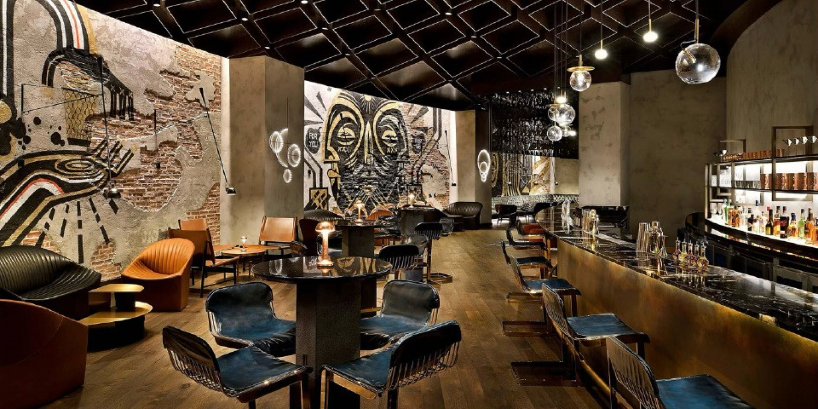 Interior of a stylish bar with modern decor, including a long bar counter with barstools, eclectic seating, wall art, exposed brick, and decorative lighting.