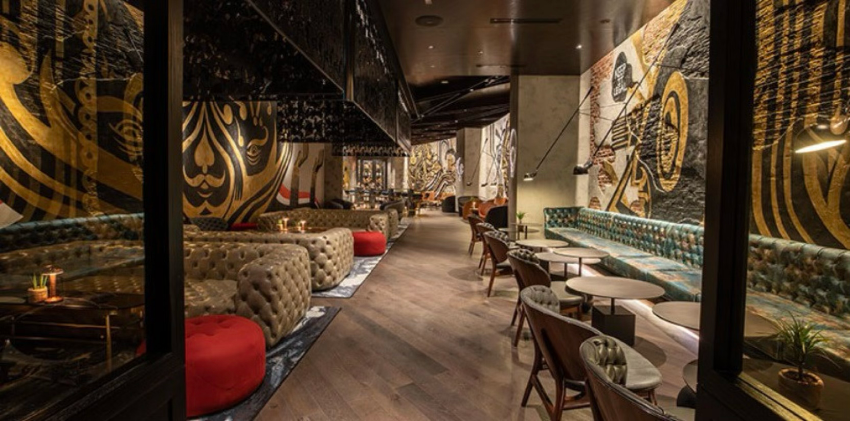 Elegant lounge with assorted seating including tufted sofas and patterned chairs, dark wood flooring, artistic wall murals in black, gold, and beige, warm ambient lighting, and small tables, creating a cozy and stylish atmosphere.
