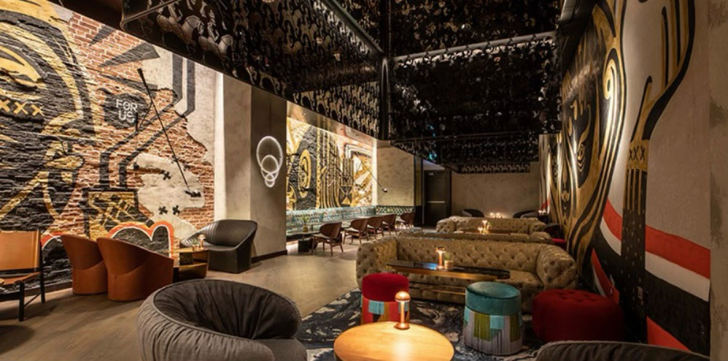 Luxurious bar or lounge with eclectic decor, featuring plush chairs, colorful curtains, exposed brick walls with graffiti art, and ambient lighting.