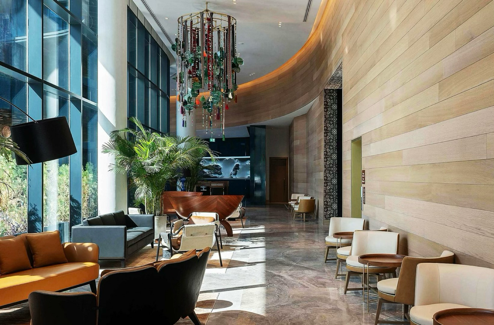 Modern hotel lobby with large floor-to-ceiling windows, stylish seating, indoor plants, a chandelier, and wooden wall panels.
