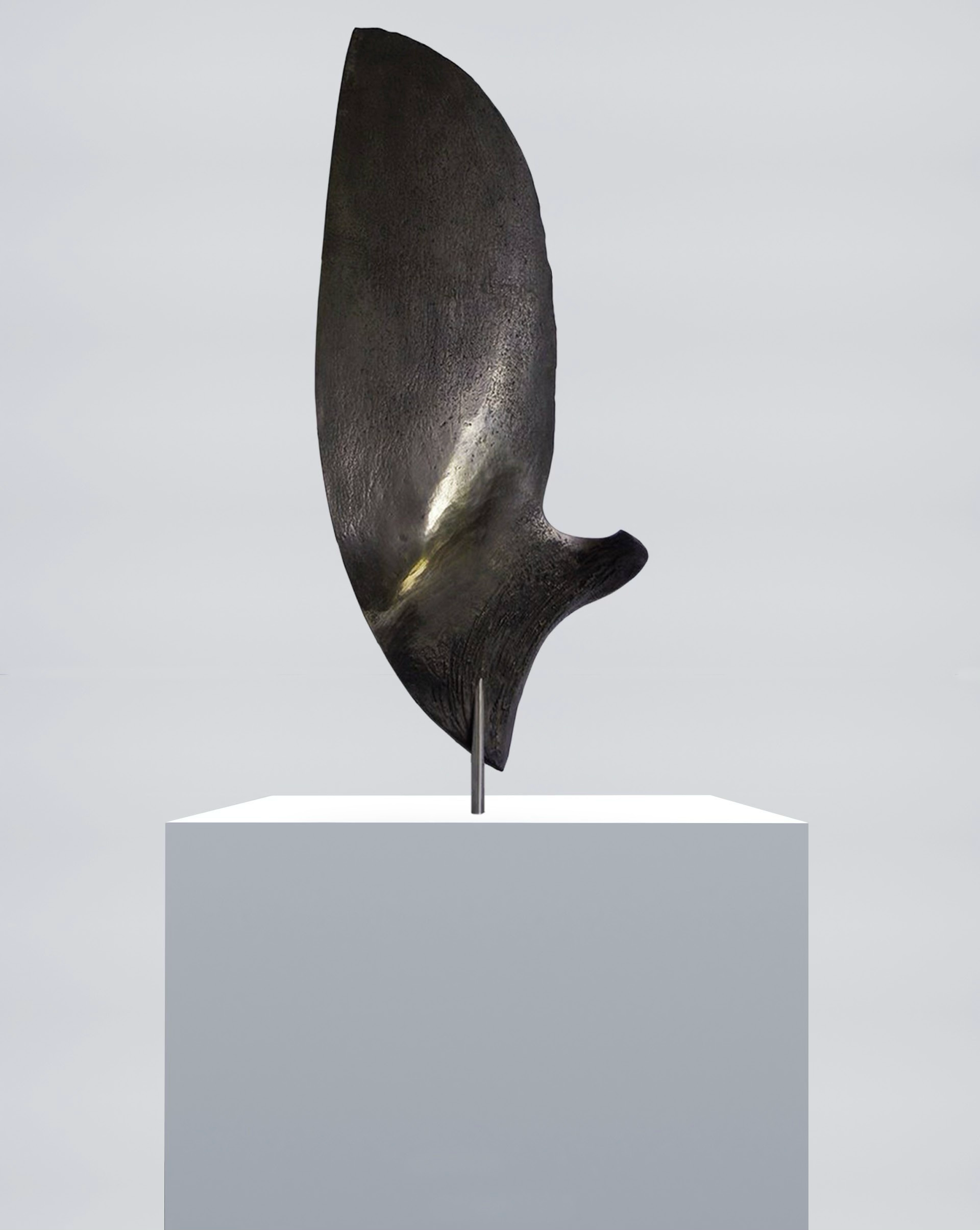 A modern sculpture resembling a whale fin displayed on a white pedestal against a neutral background.