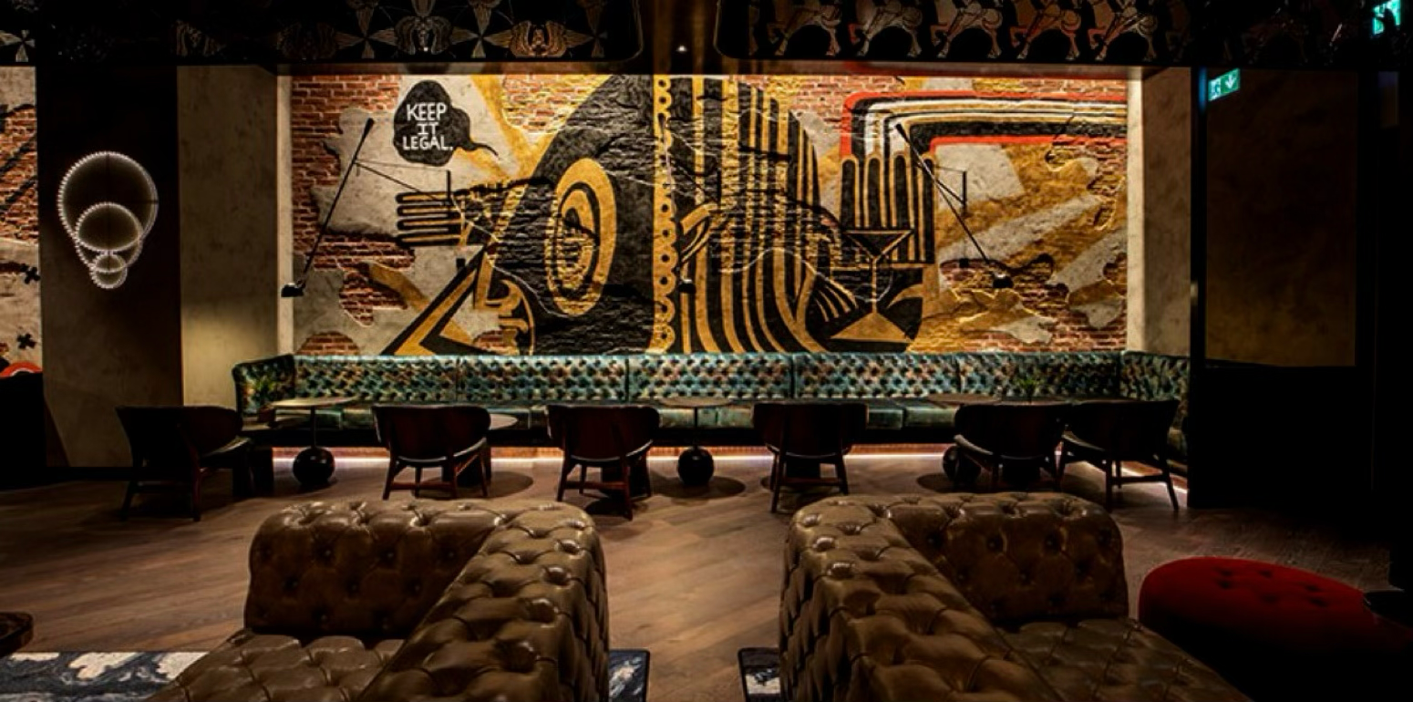 Interior of a stylish lounge or bar with modern decor, including a large colorful mural on the back wall, a long patterned sofa, and leather armchairs.