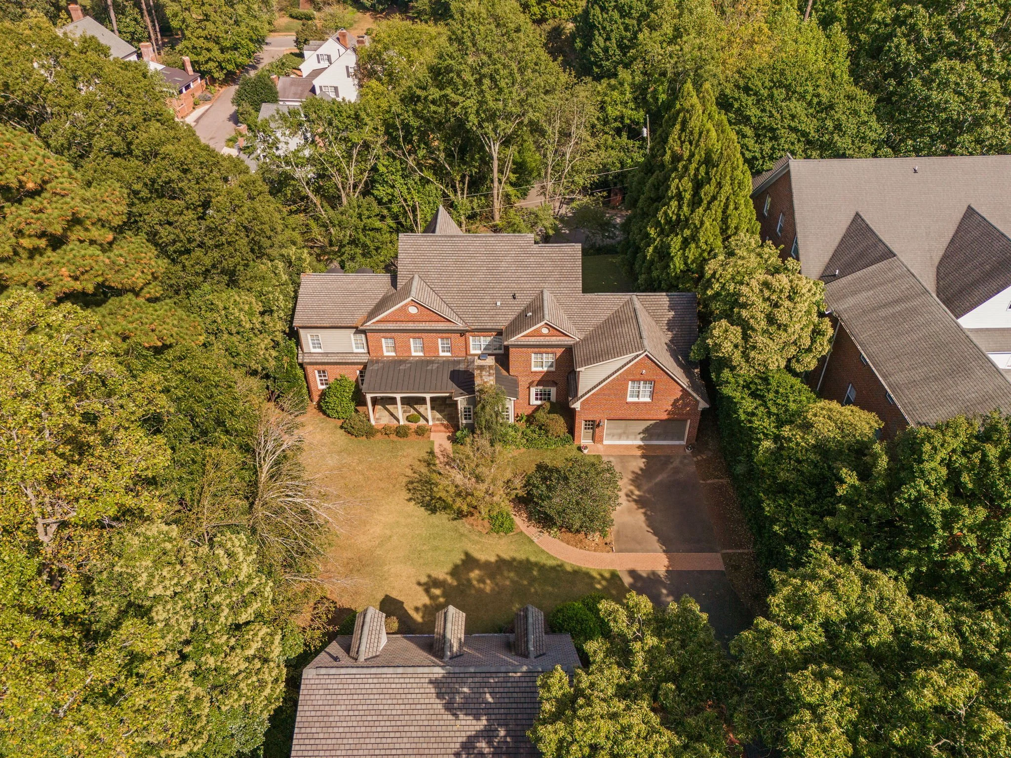Aerial view of a two-story brick house with a front porch, surrounded by a backyard with grass, trees, and shrubs, and neighboring houses in a suburban neighborhood.