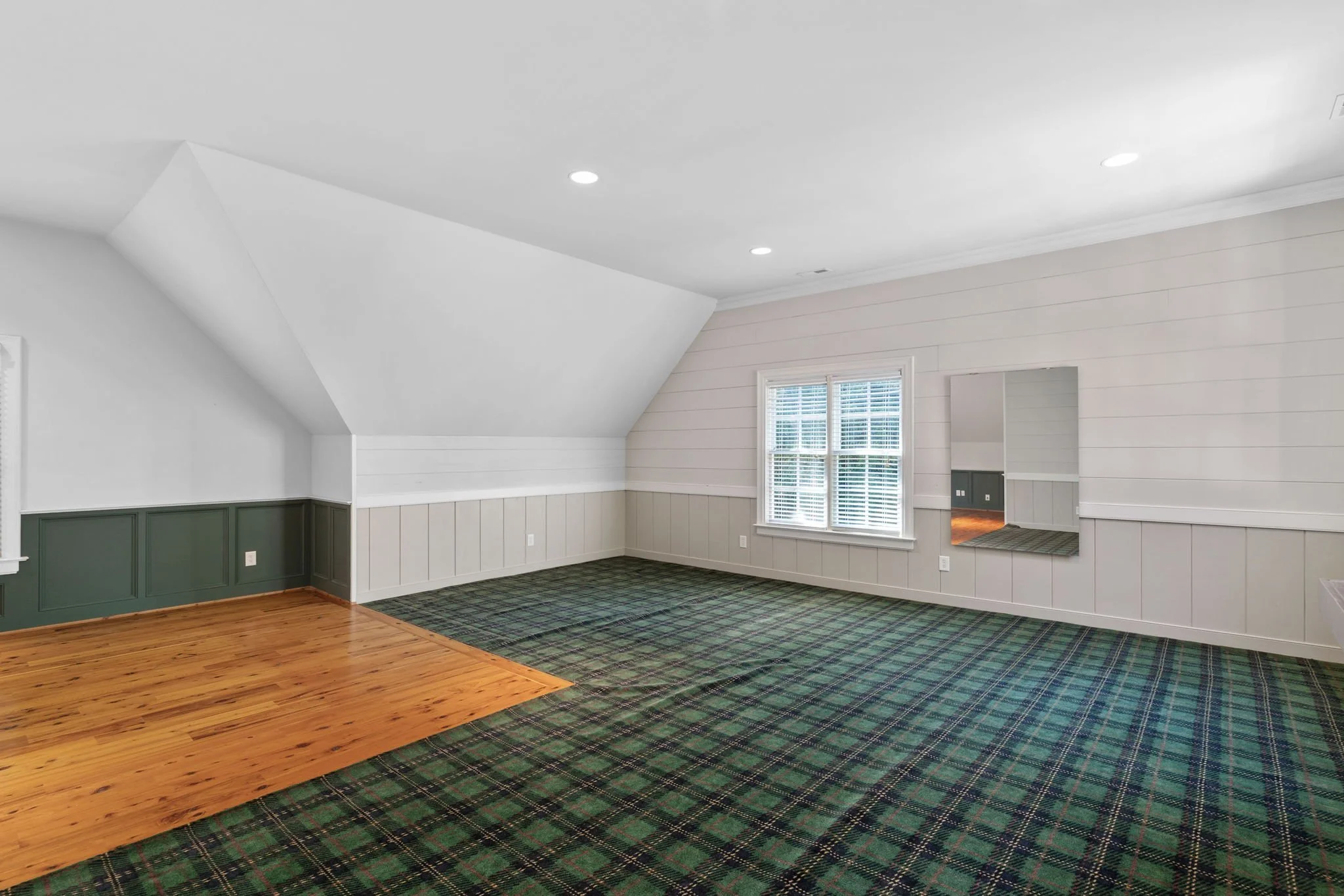Empty room with white walls, a window with blinds, and a mix of wood and checkered carpet flooring.