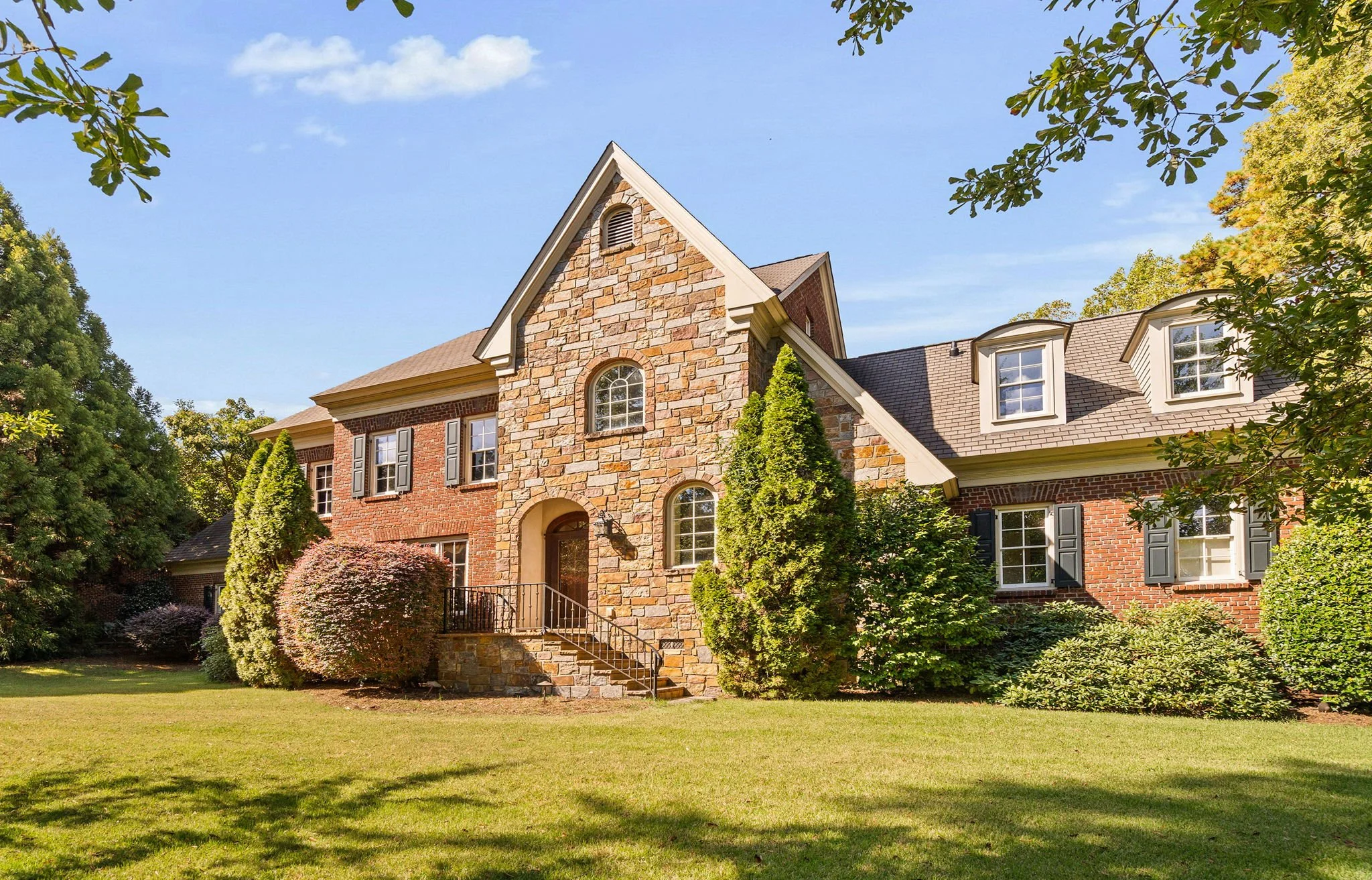 A large brick and stone house with multiple windows, surrounded by well-maintained landscaping and trees under a blue sky.