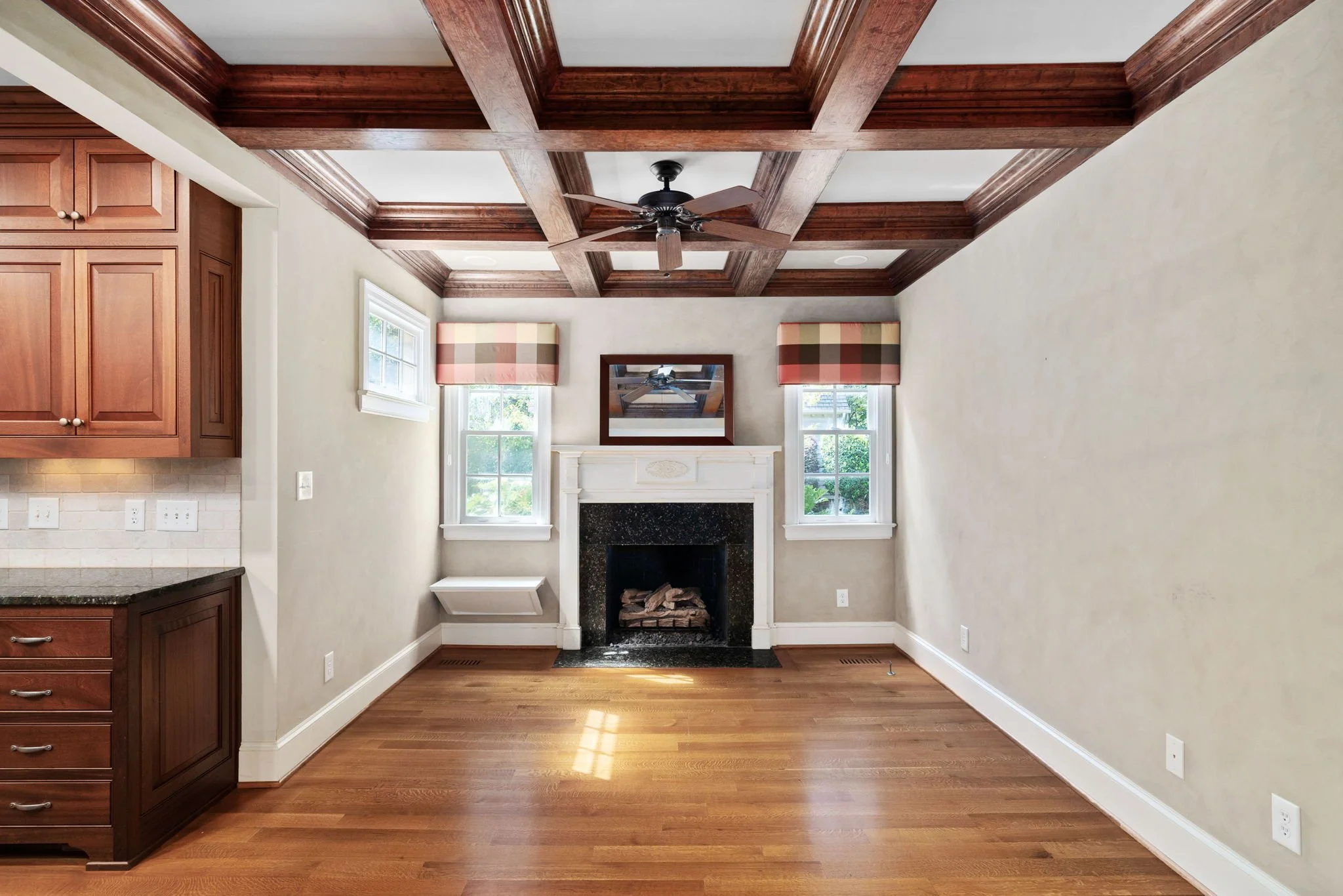 Living room with hardwood floors, beige walls, and a white fireplace with black hearth. Two windows with plaid valances, a mirror above the fireplace, and a ceiling fan with dark blades. Part of a kitchen with wooden cabinets is visible on the left.