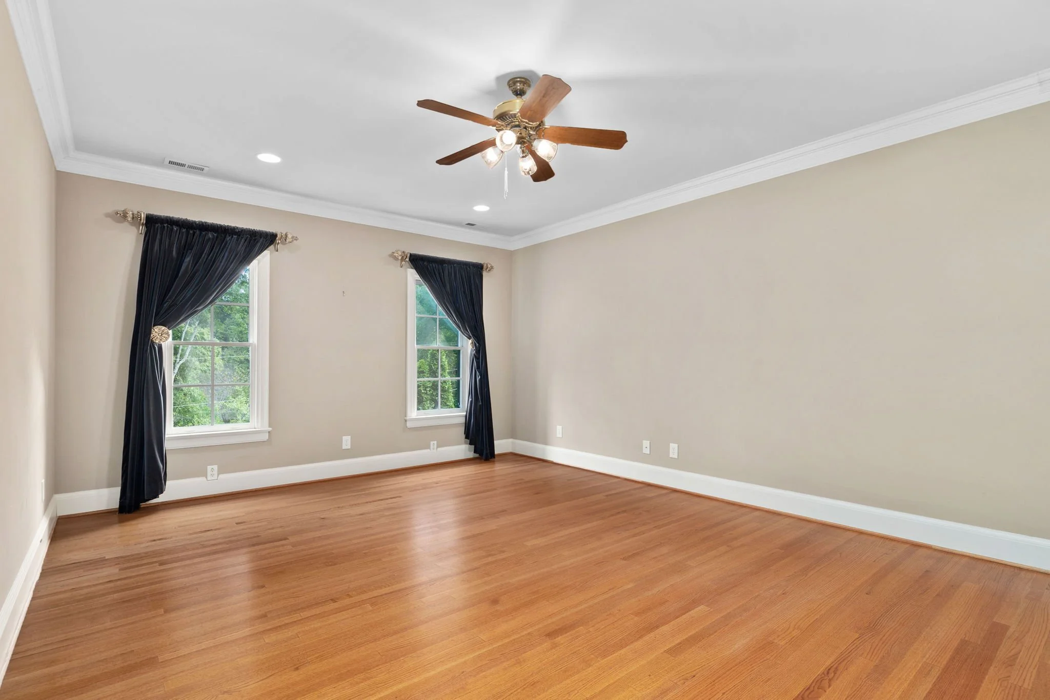 Empty living room with beige walls, hardwood floors, two windows with dark curtains, white crown molding, and a ceiling fan with lights.