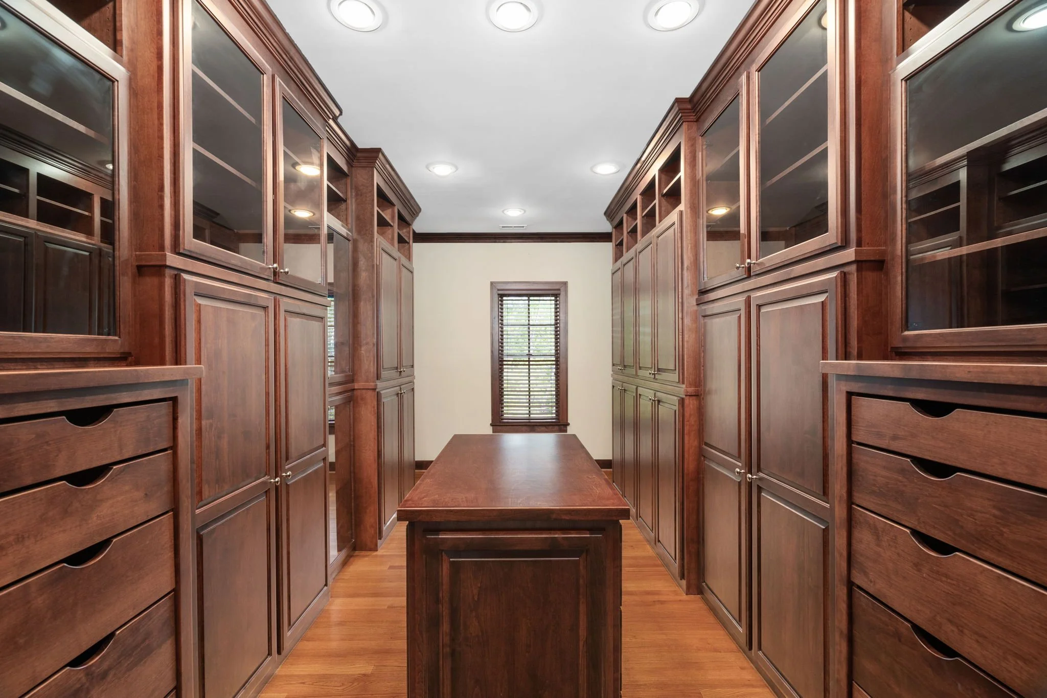 Wooden walk-in closet with built-in cabinets and drawers, a central table, hardwood floors, and a small window with blinds.