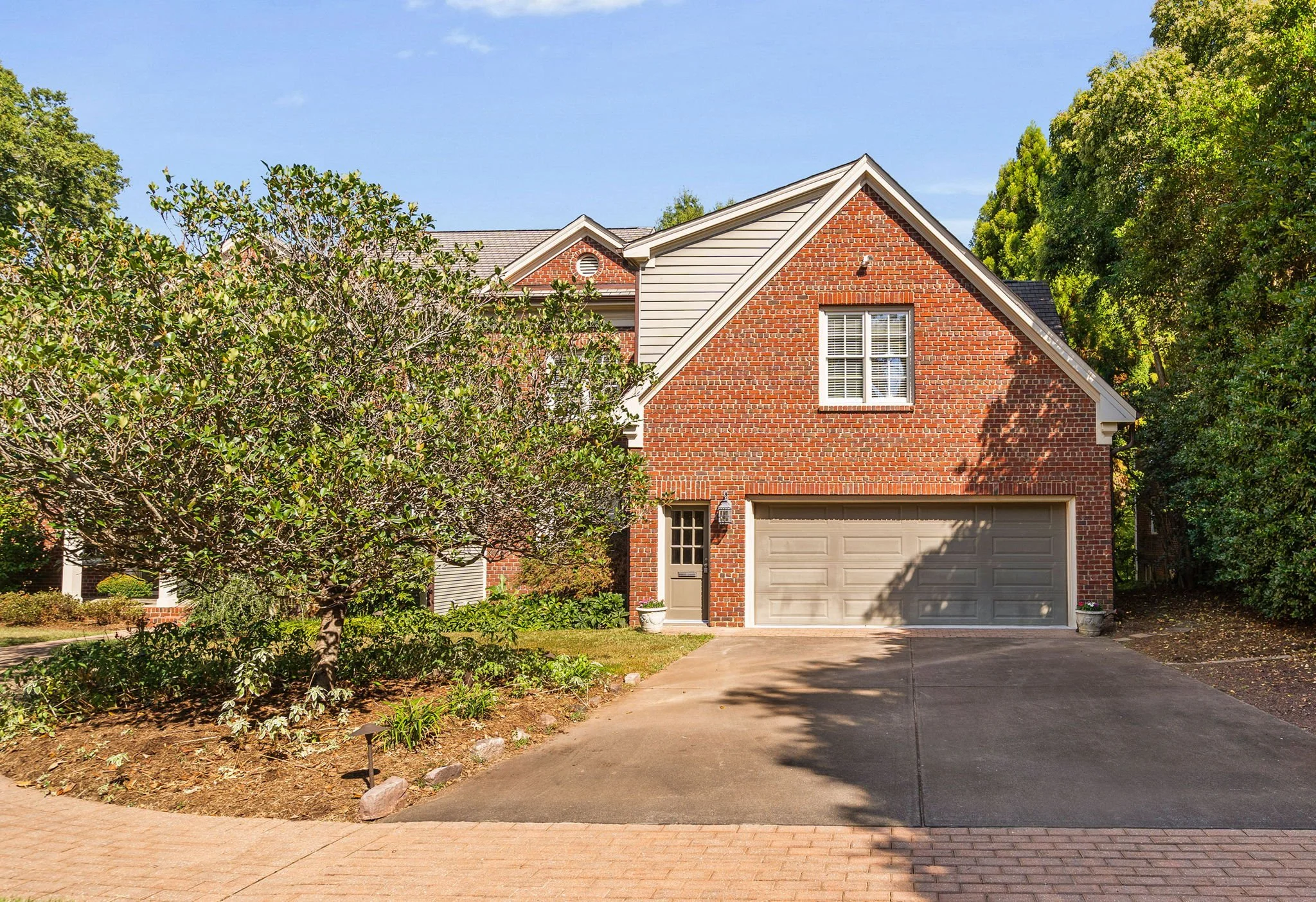 Front view of a two-story brick house with a closed garage and a small side door, surrounded by trees and greenery under a clear blue sky.
