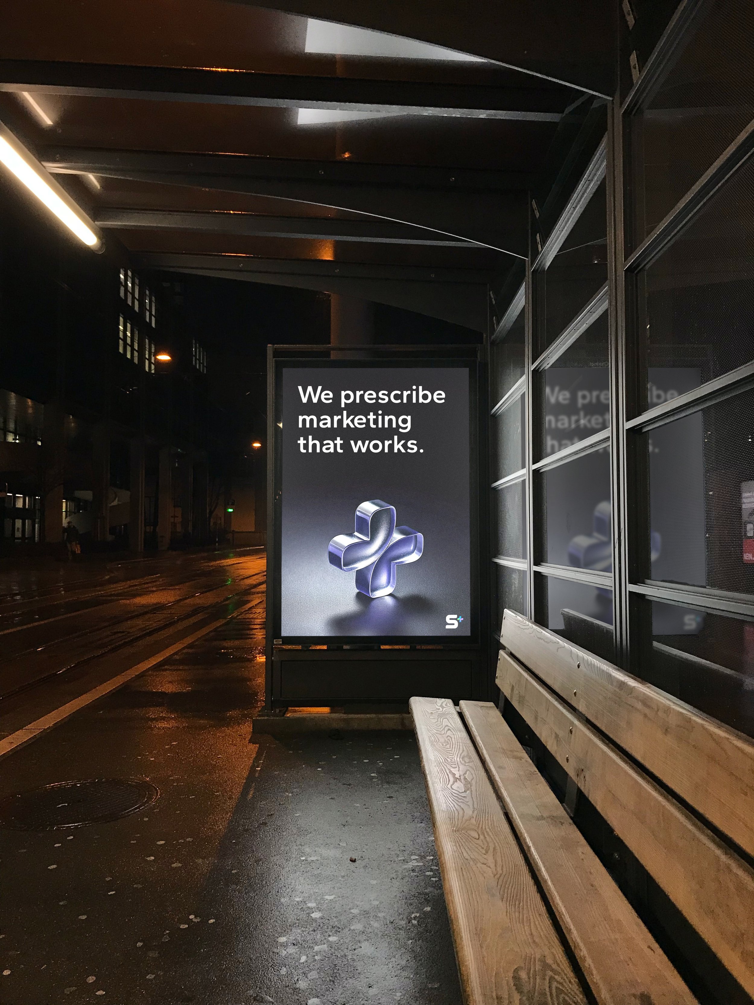Empty bus stop with a digital advertisement displaying the message 'We prescribe marketing that works' next to a wooden bench at night on a wet street.