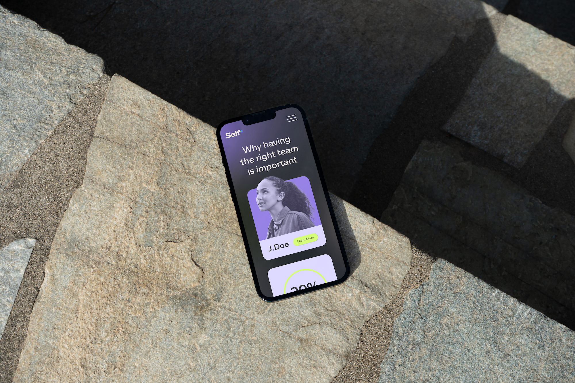 Smartphone displaying a website article titled 'Why having the right team is important,' with a photo of a smiling woman and buttons labeled 'Learn More' and a percentage indicator, resting on a stone surface.