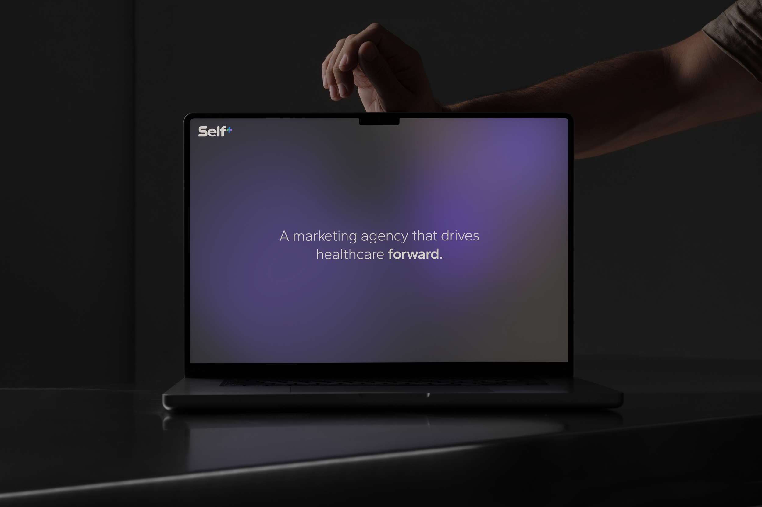 A laptop displaying a website for Self, a marketing agency, with the tagline "A marketing agency that drives healthcare forward," set against a dark background with a person's hand hovering above.