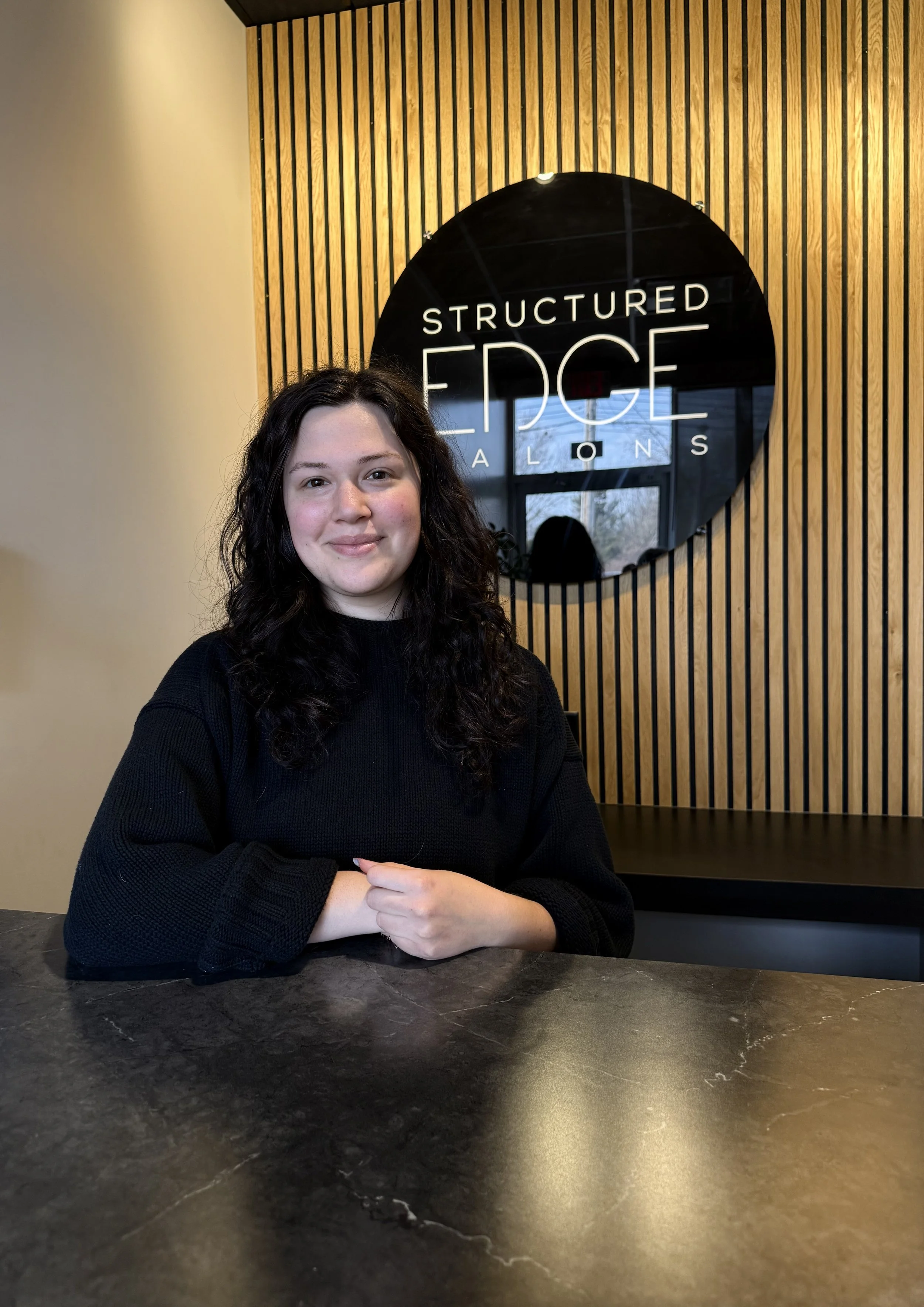 Smiling person at reception desk of Structured Edge Salons, with wooden paneling and potted plants in background.