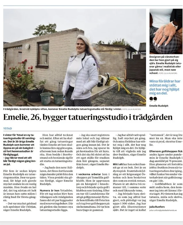 En kvinna står utomhus framför ett trädgårdsavgränsning, bär en vit jacka och ler, med en byggnad i bakgrunden samt träd och buskar.