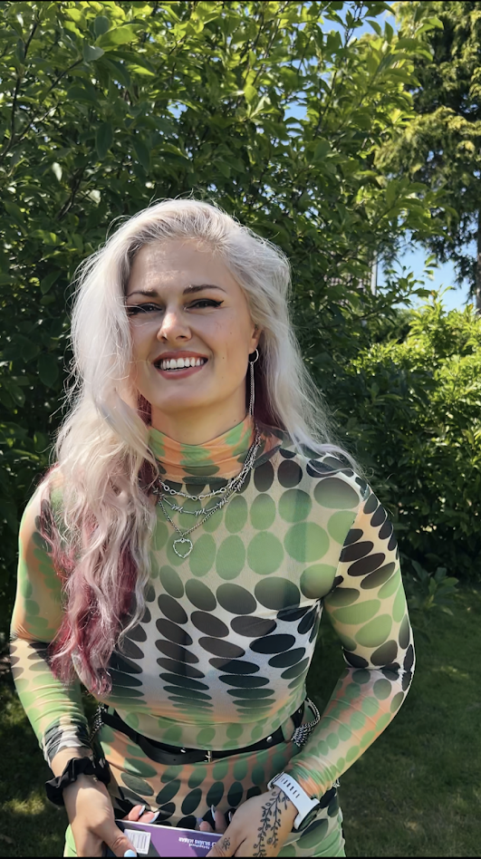 Kvinna med långt blont hår och grön mönstrad tröja står utomhus framför grönska, ler mot kameran.